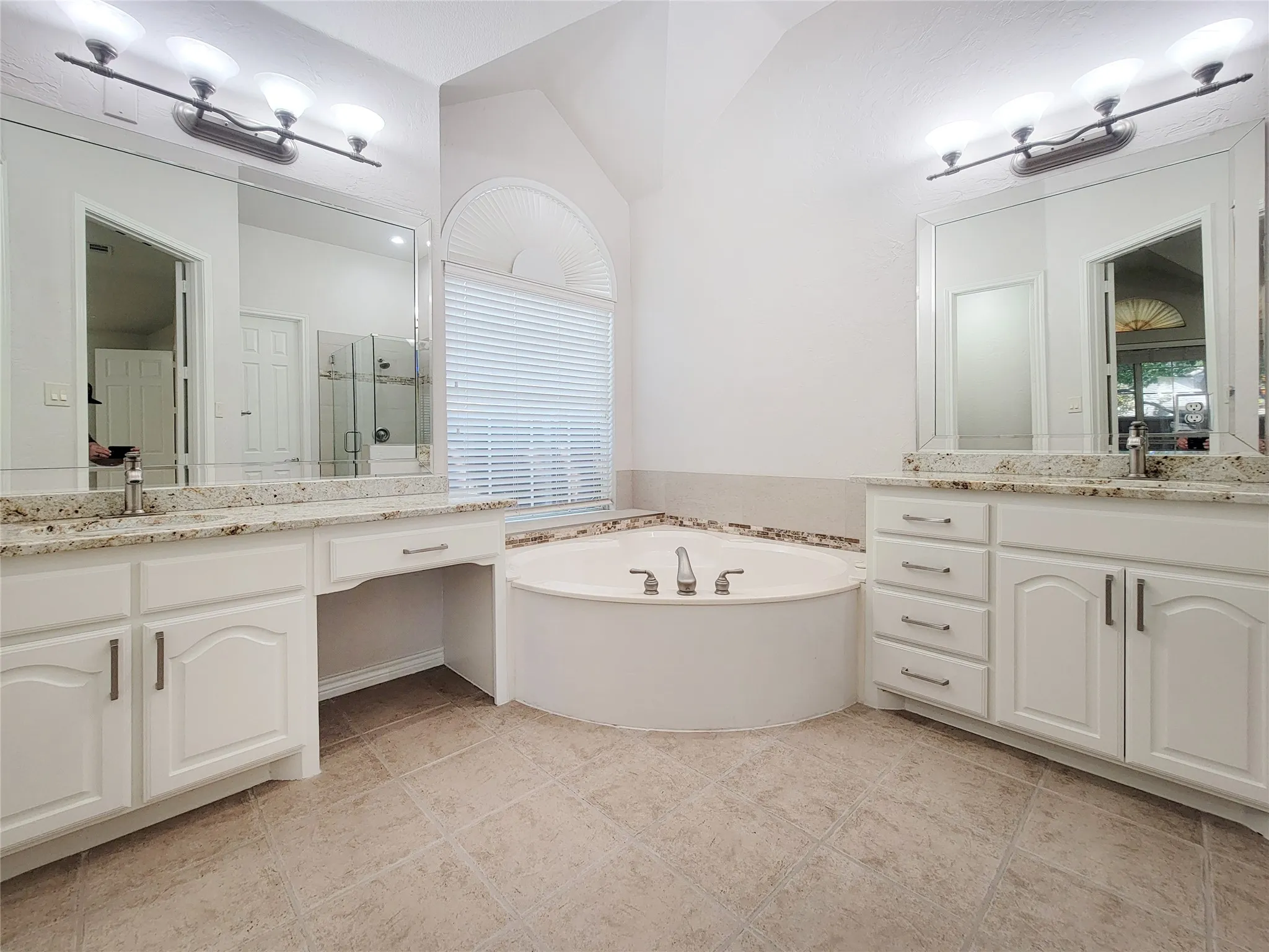 Dual vanity with granite countertops, integrated drop-in soaking tub, arched window, and tile flooring