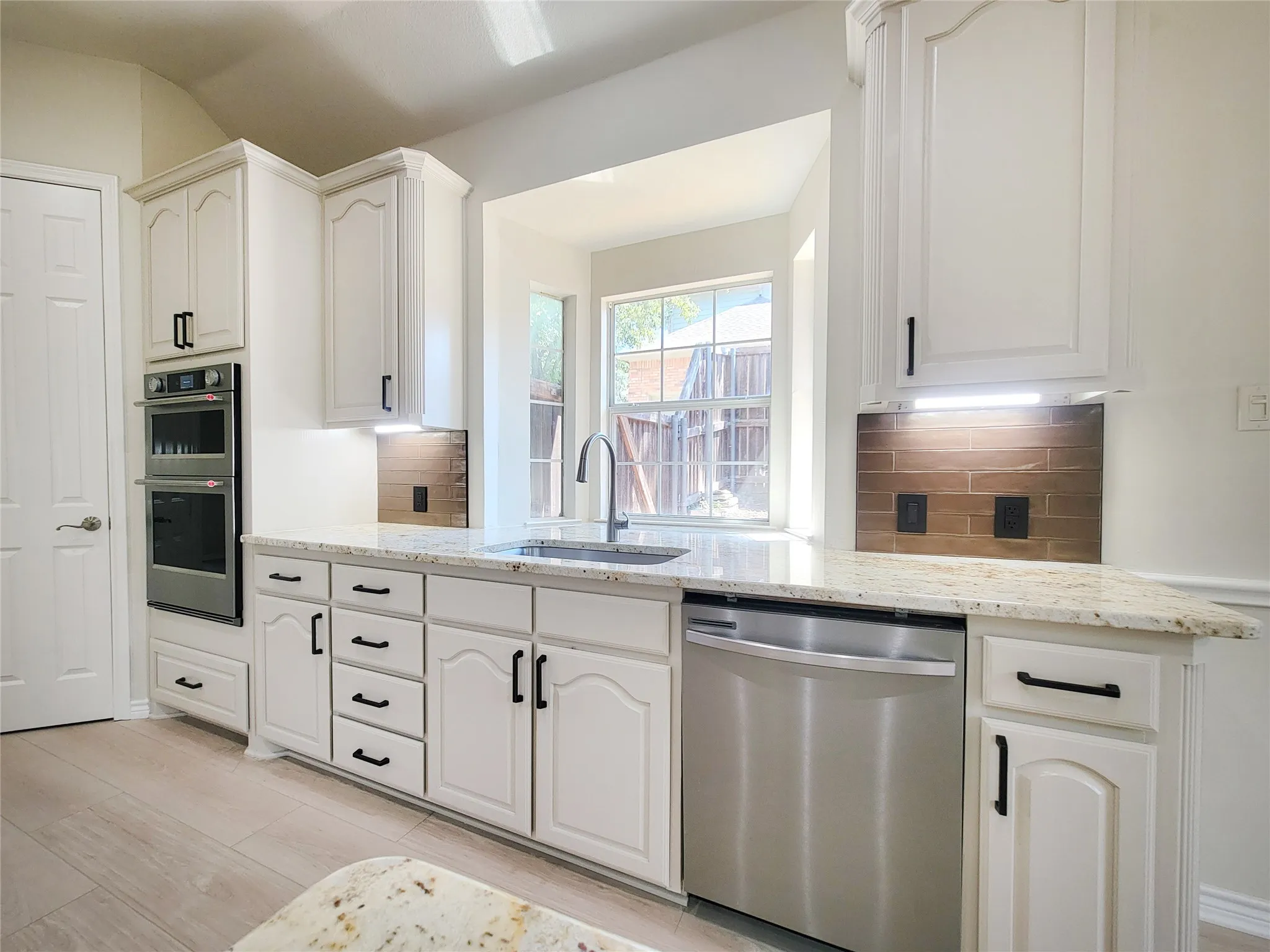 White cabinetry with black hardware, stainless steel built-in double oven, stainless steel dishwasher, speckled light-toned countertops, and a built-in sink with a gooseneck faucet