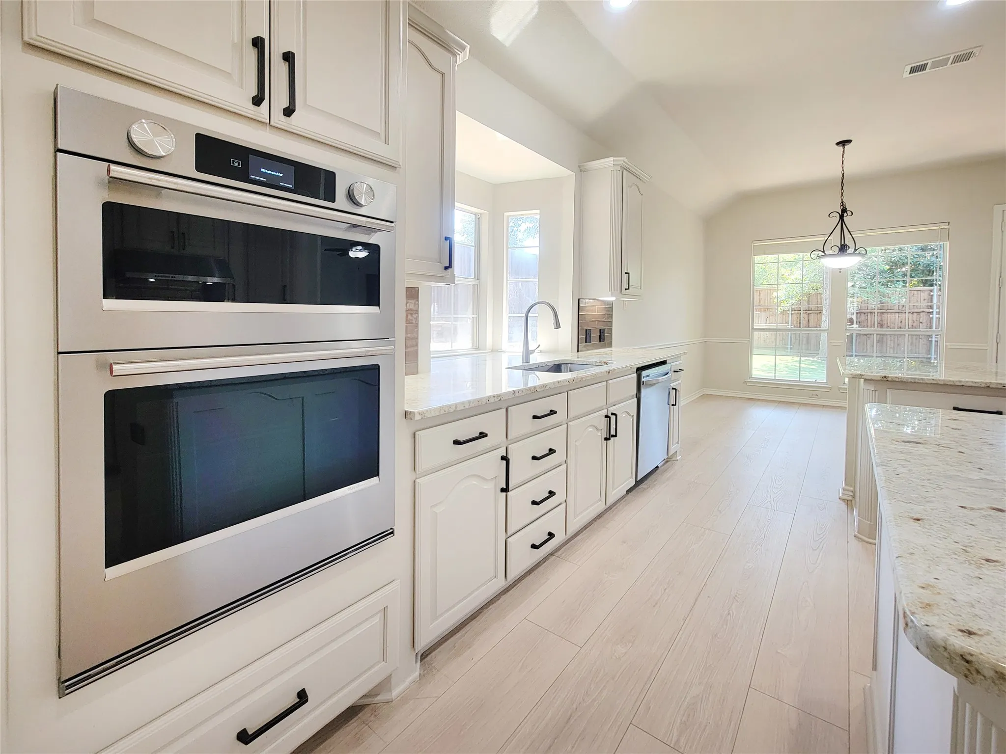 Stainless steel built-in double ovens, white cabinetry with black hardware, light-toned wood-finish flooring, and white countertops