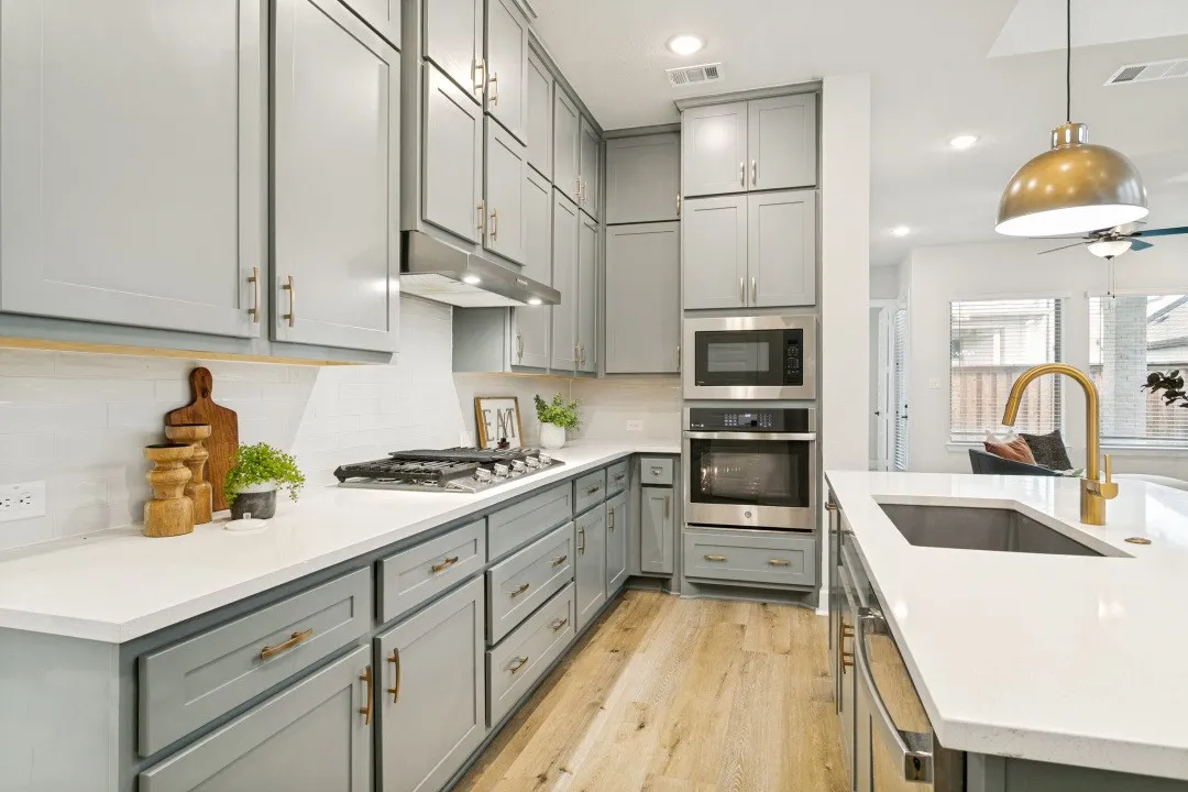 Contemporary kitchen featuring light gray cabinetry with gold-toned hardware, stainless steel appliances, white countertops, and wood-finish flooring