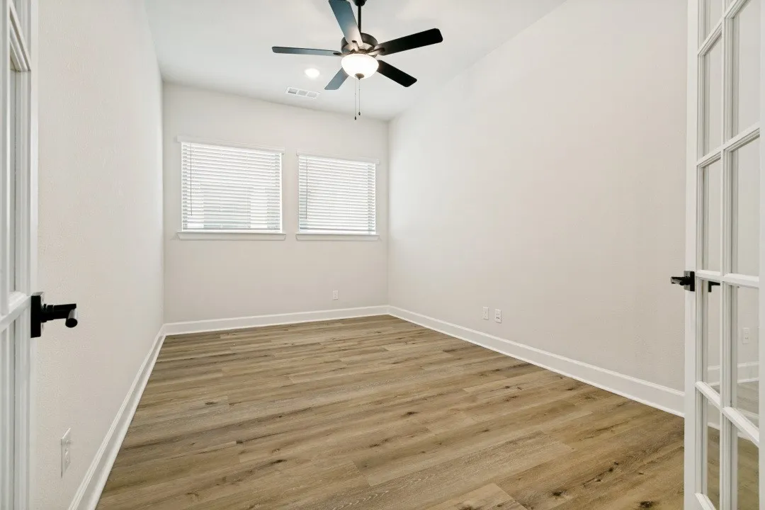 Room featuring wood-finish flooring, two windows with blinds, a ceiling fan with integrated lighting, and French doors with black hardware