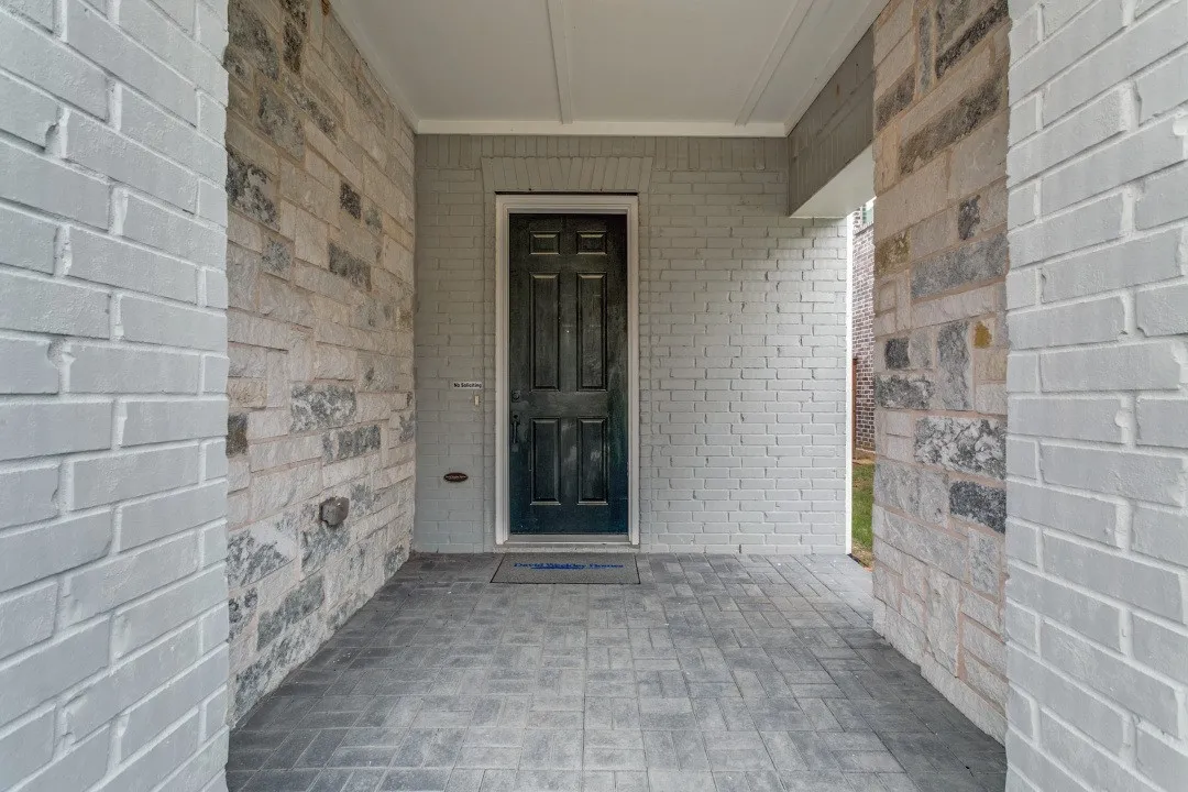 Covered entryway featuring a dark paneled door, gray painted brick walls, natural stone accents, and a stamped concrete floor