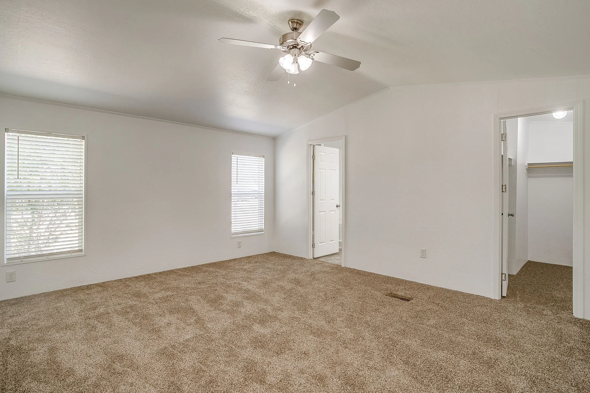 Spacious bedroom with neutral carpet flooring, ceiling fan with integrated lighting, abundant natural light from dual windows with plantation shutters, and convenient access to an ensuite bathroom