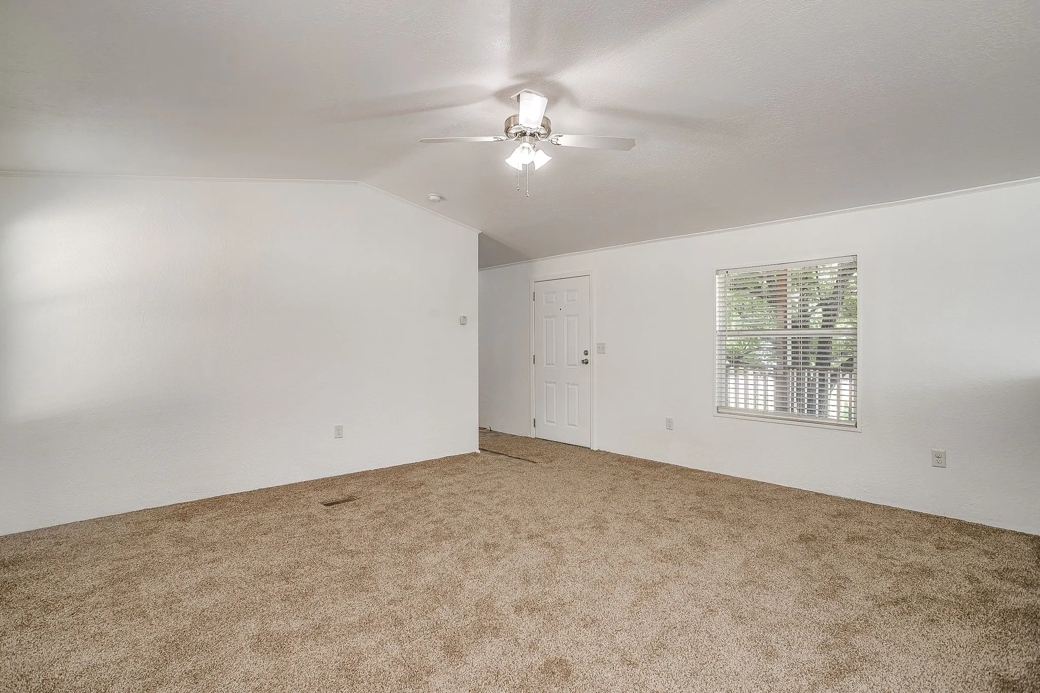 Spacious carpeted room with neutral wall finishes, ceiling fan with integrated lighting, and abundant natural light from a large window with plantation shutters