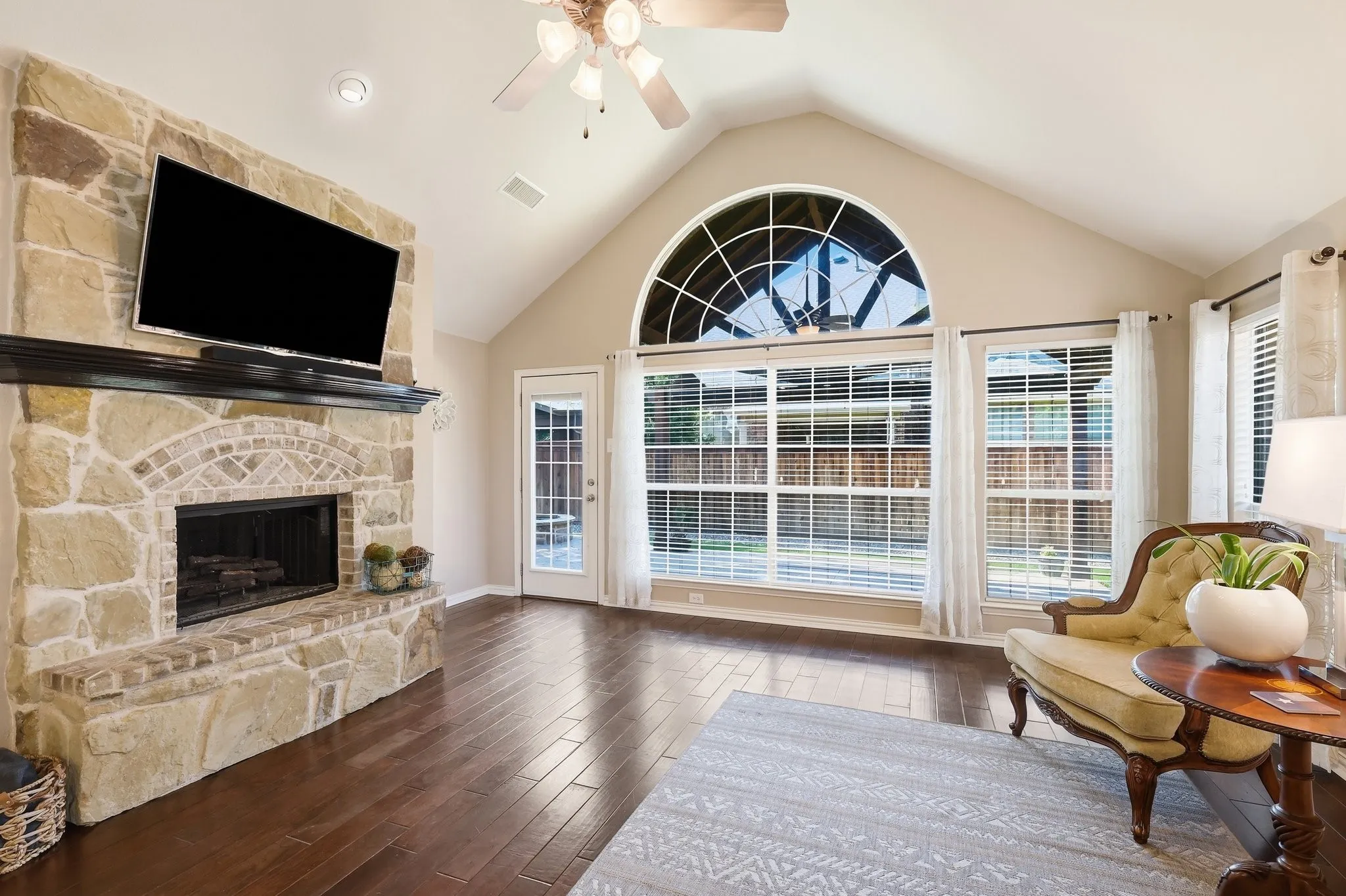 Cathedral ceiling with a ceiling fan, arched transom window, wood-finish flooring, brick fireplace surround, and multiple windows with blinds