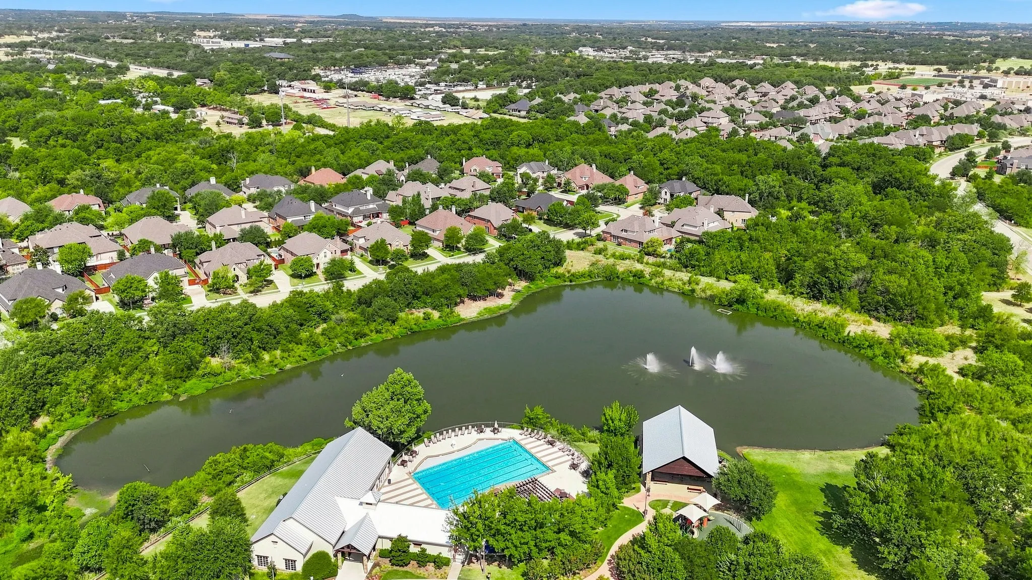 Expansive community pond with water features, surrounded by mature trees, and featuring a swimming pool, and pavilion