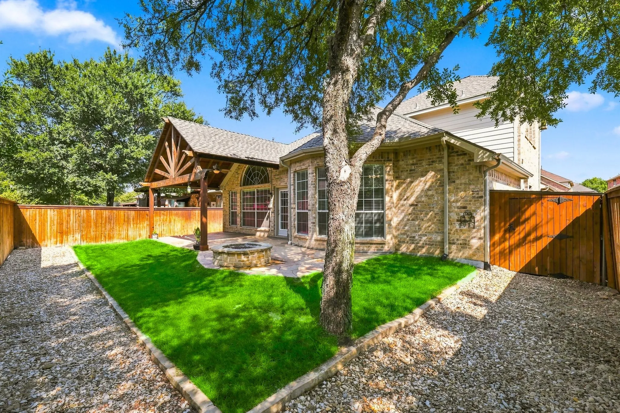 Brick exterior with multiple windows, covered patio, stone fire pit, and a fenced yard with lawn and gravel areas