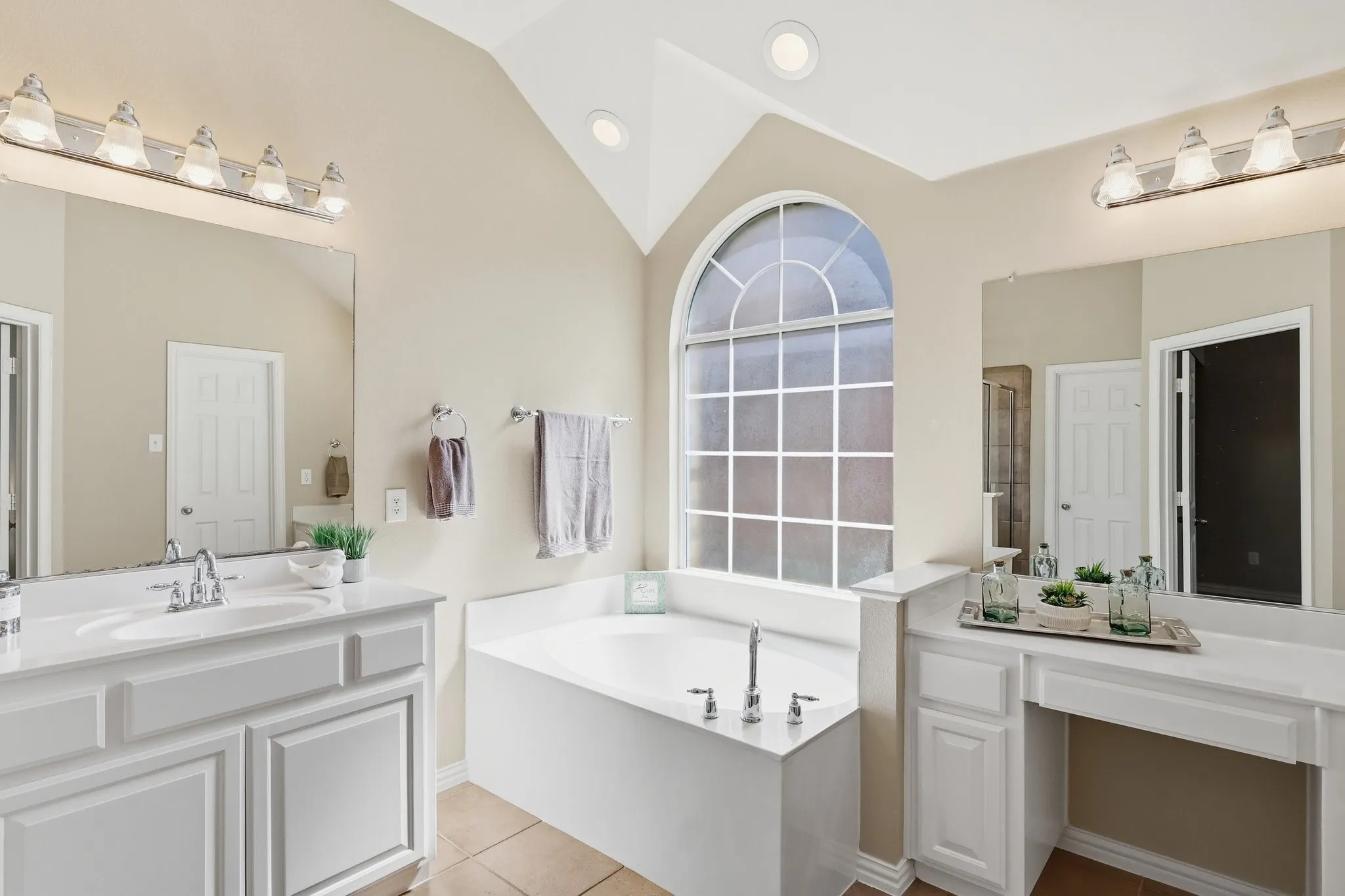 Vaulted ceiling with recessed lighting, arched window with grid, built-in soaking tub, dual vanities with white cabinetry, and tile flooring