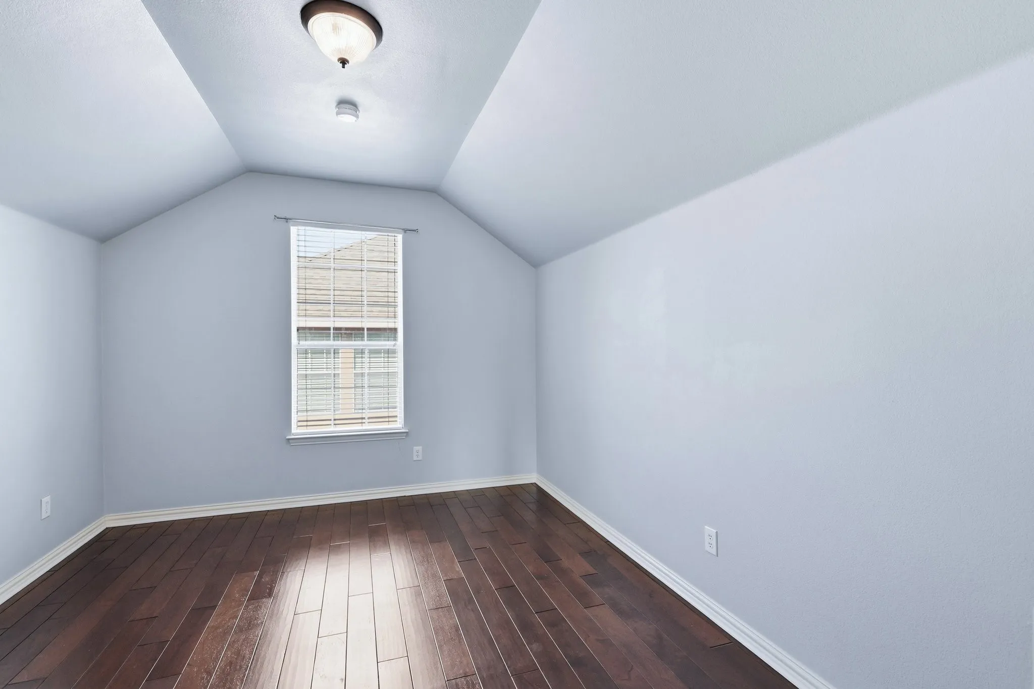 Vaulted ceiling with a flush-mount light fixture, a single window with blinds, and rich wood-finish flooring