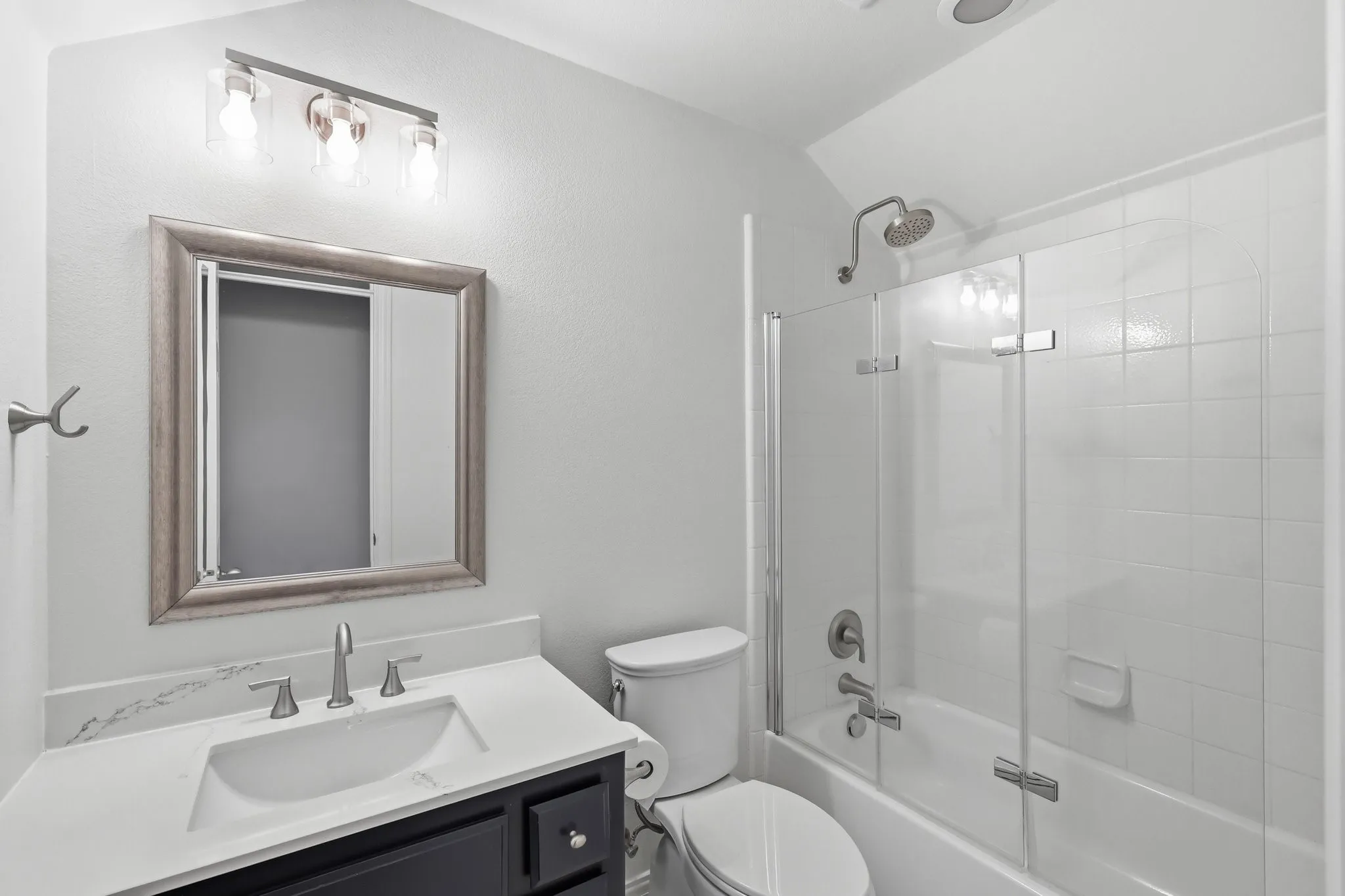 Vanity with built-in sink, framed mirror, three-light vanity bar, white countertop, and glass-enclosed shower-tub combination