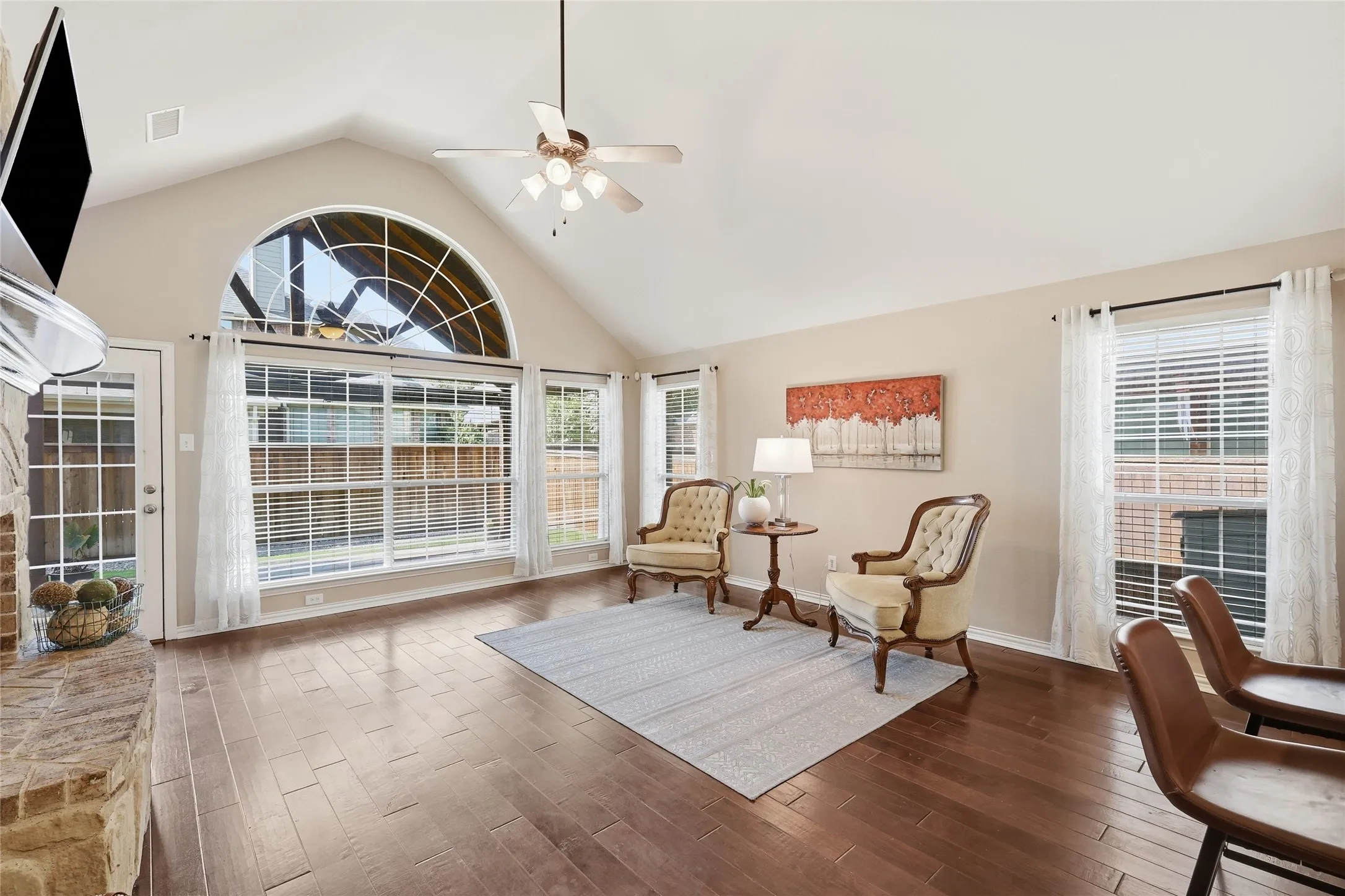 Cathedral ceiling with a ceiling fan, arched transom window, wood-finish flooring, brick fireplace surround, and multiple windows with blinds