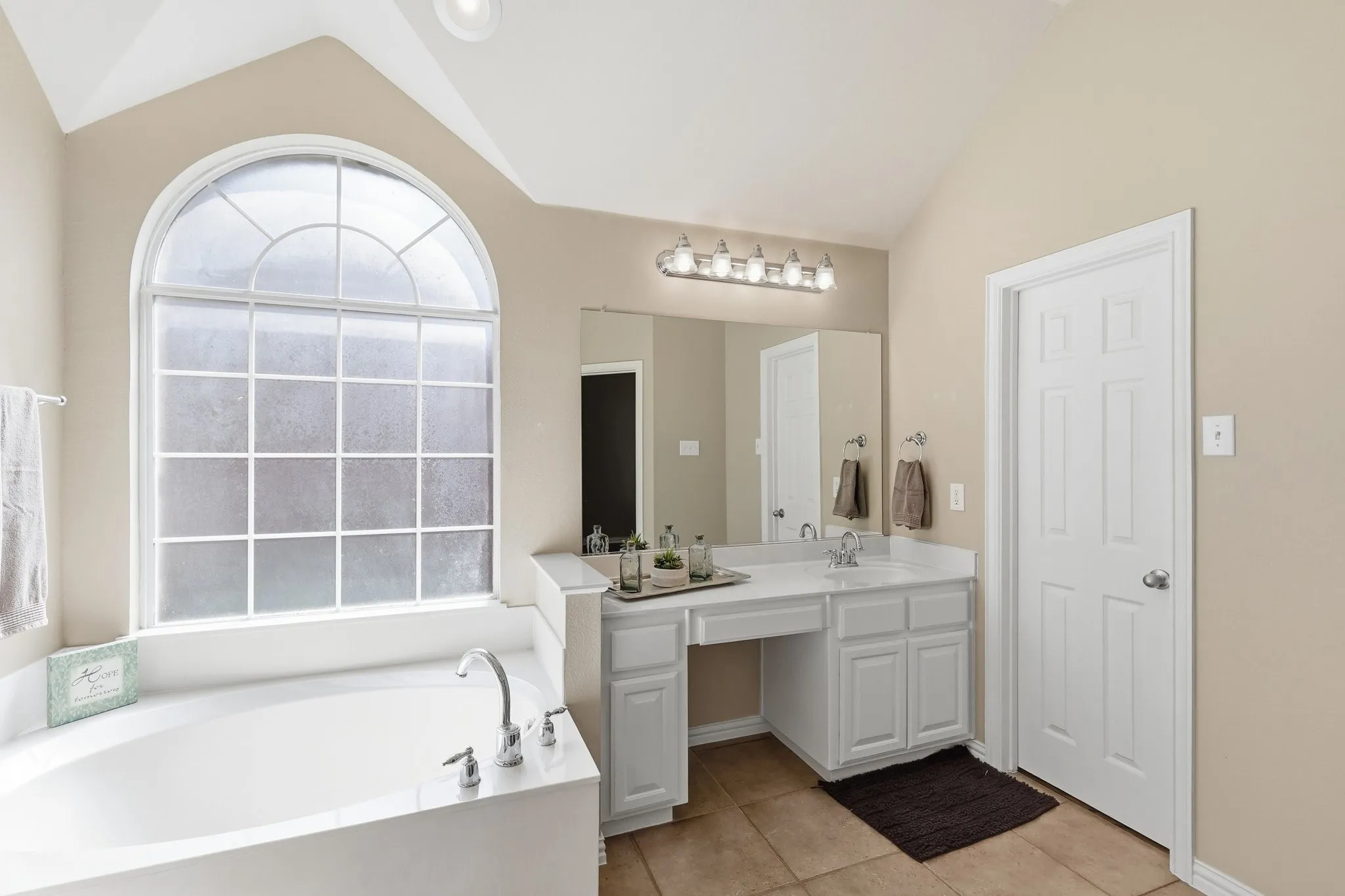 Arched window with privacy glass, built-in soaking tub with deck-mounted fixtures, dual vanity with undermount sinks, and tiled flooring