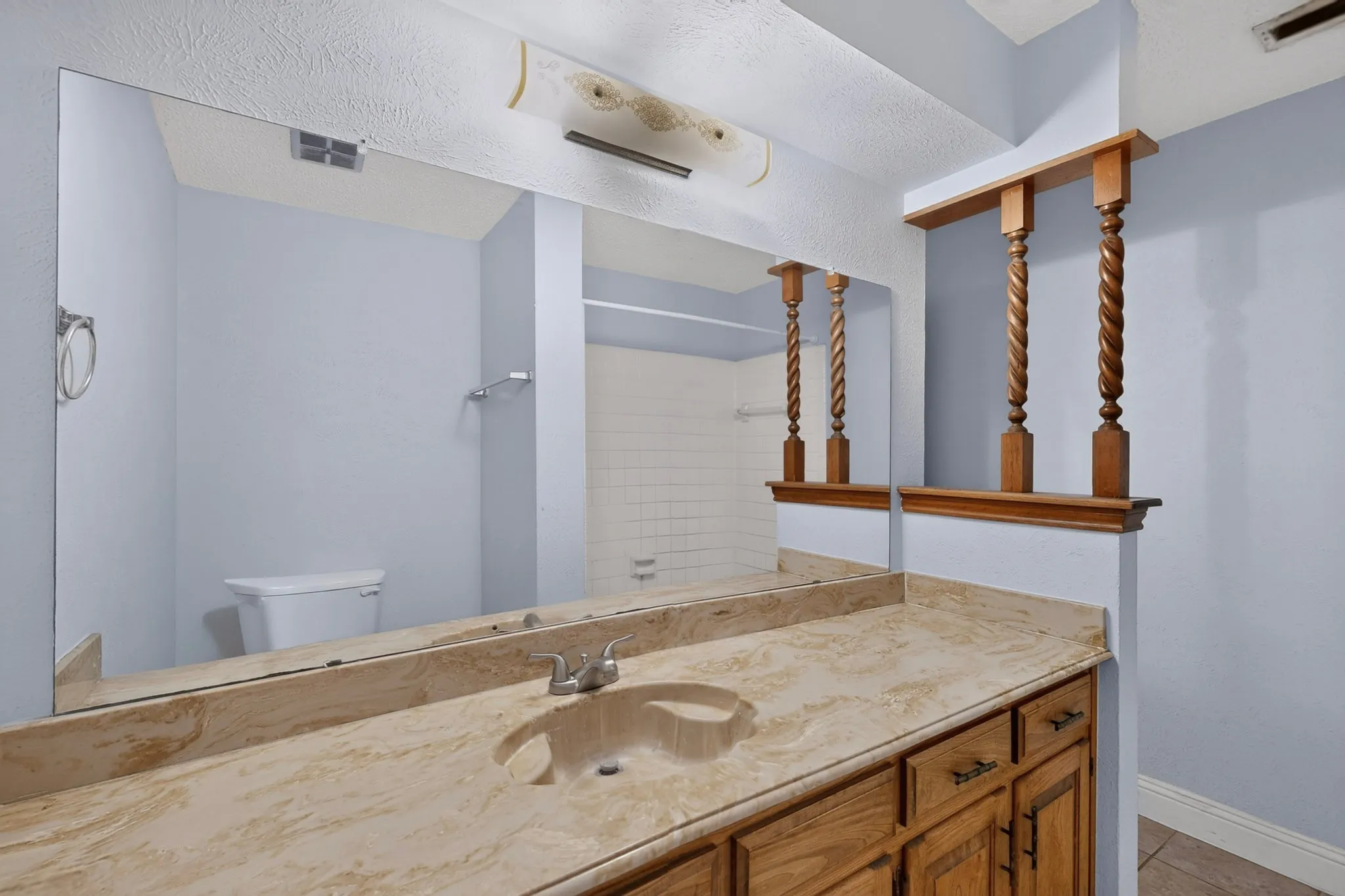 Full-width vanity mirror, integrated vanity sink, light-toned stone vanity countertop, wood-finish vanity cabinetry with dark hardware, and decorative wood pillars