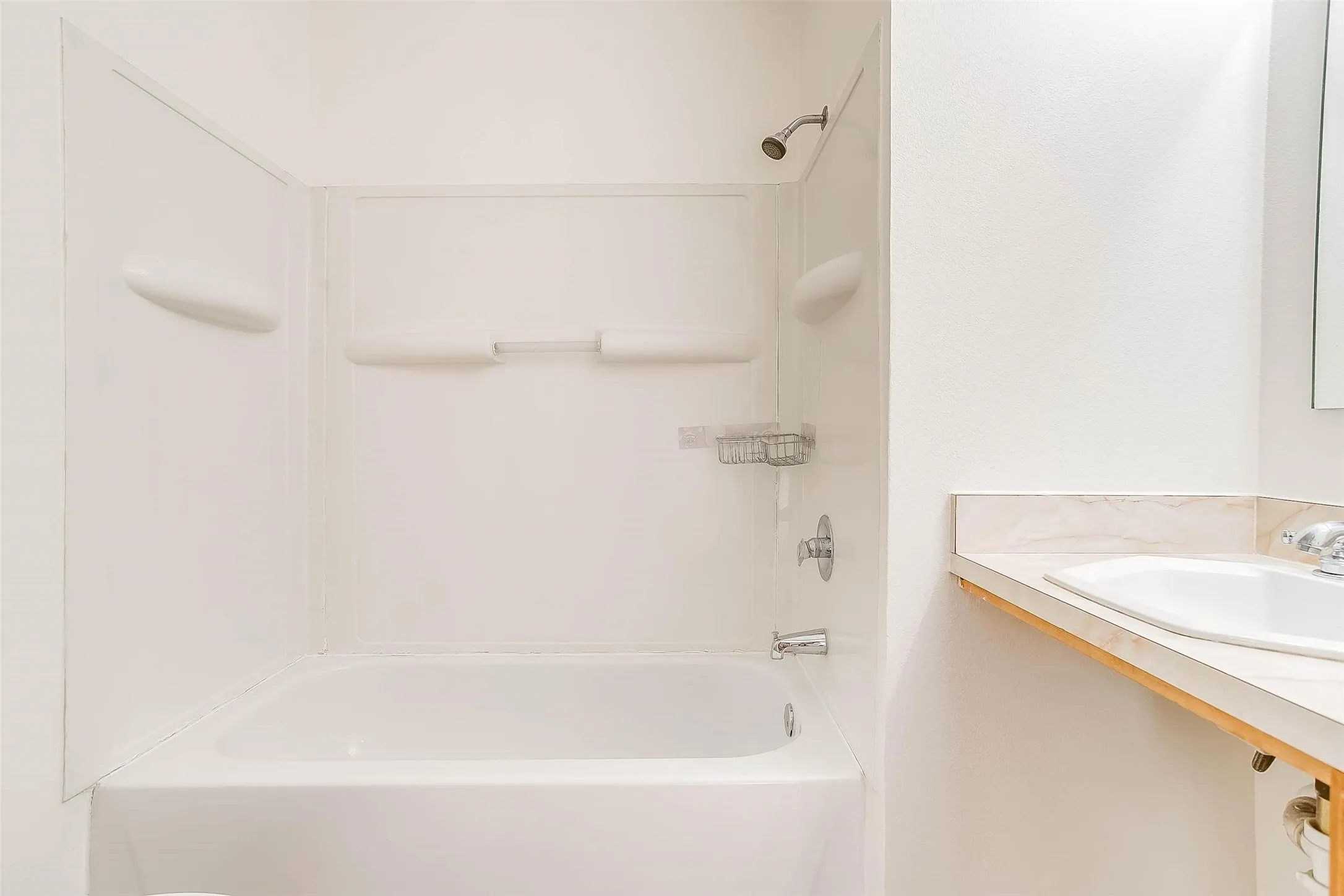 Bathtub and shower combination with chrome fixtures, integrated shelving, and a vanity with an integrated sink and light-toned countertop