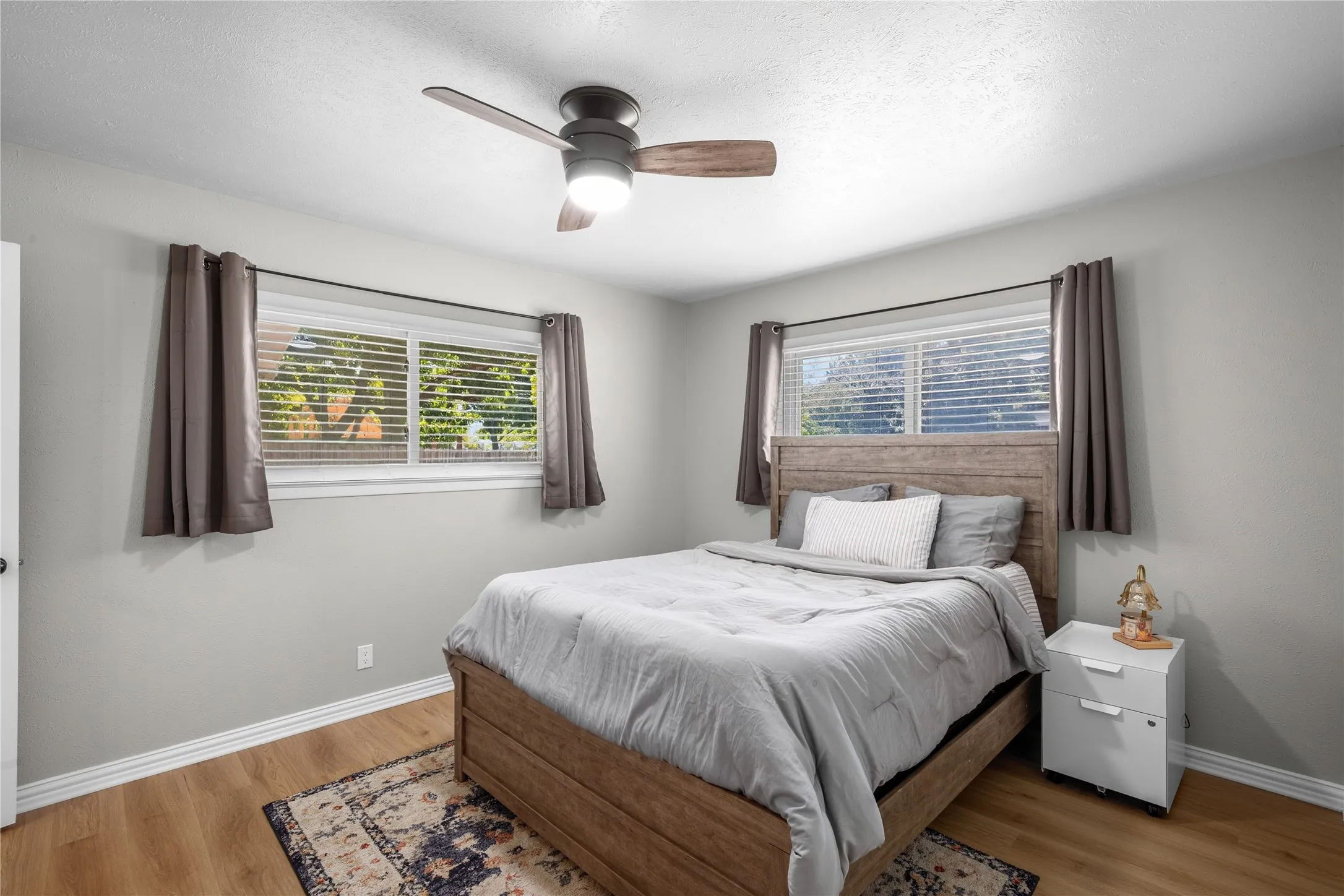 Light wood-finish flooring, two large windows with blinds, ceiling fan with integrated lighting, and neutral wall paint