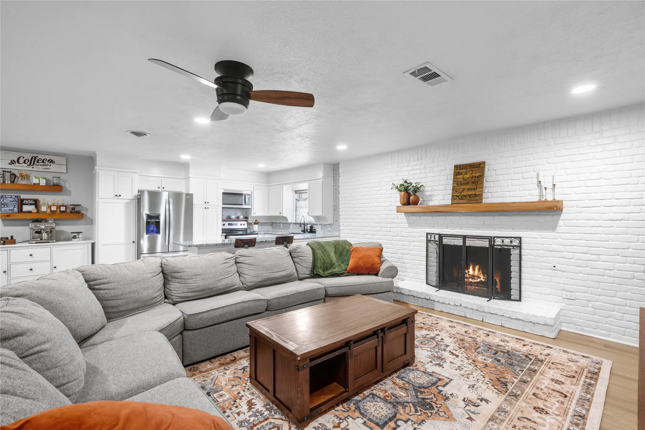 White brick fireplace with a wood mantle and hearth, recessed lighting, wood-finish flooring, a ceiling fan, and white cabinetry