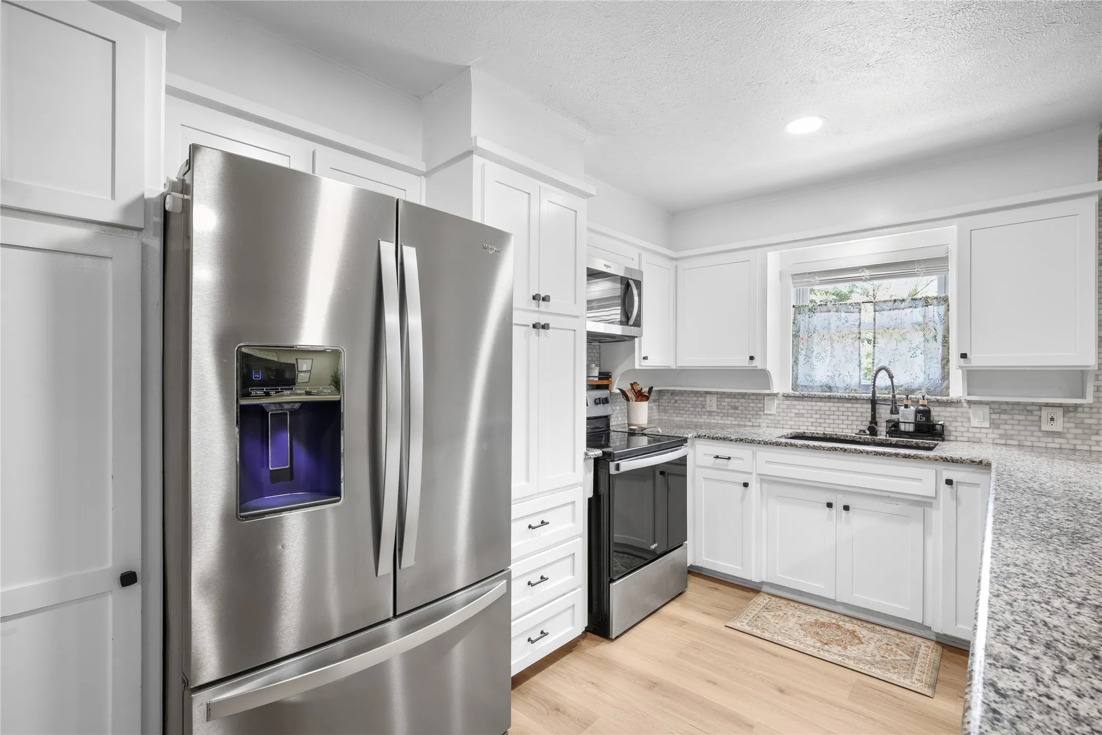Stainless steel French door refrigerator, white shaker-style cabinetry, granite countertops, subway tile backsplash, and light wood-finish flooring