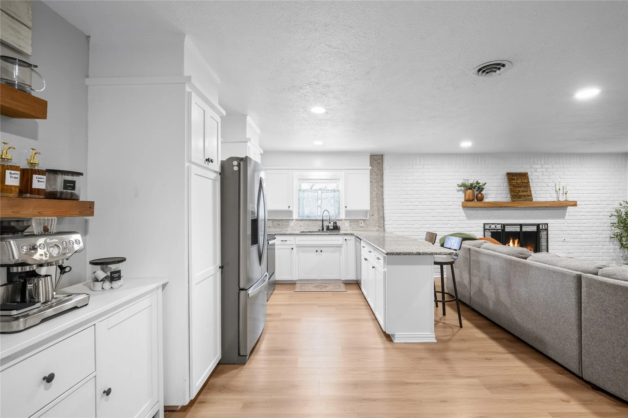 Open-concept living space featuring a white brick fireplace, wood-finish flooring, recessed lighting, and a kitchen island with stone countertops