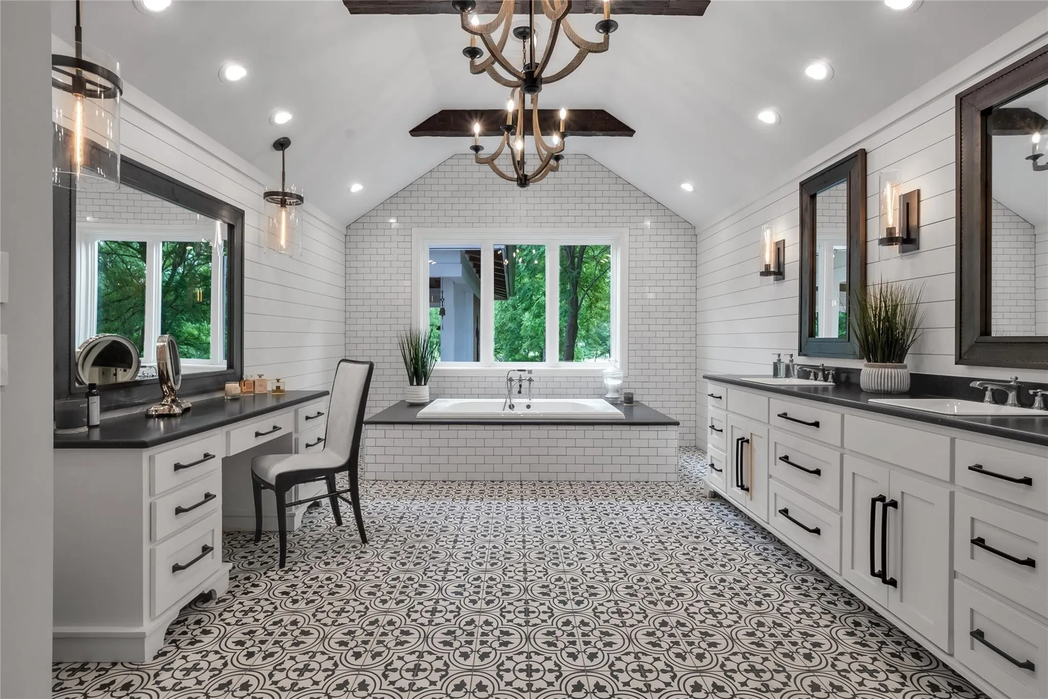 Vaulted ceiling with exposed wood beams, patterned floor tile, built-in soaking tub with subway tile surround, dual vanity with undermount sinks and dark countertops, and a dedicated vanity area with storage