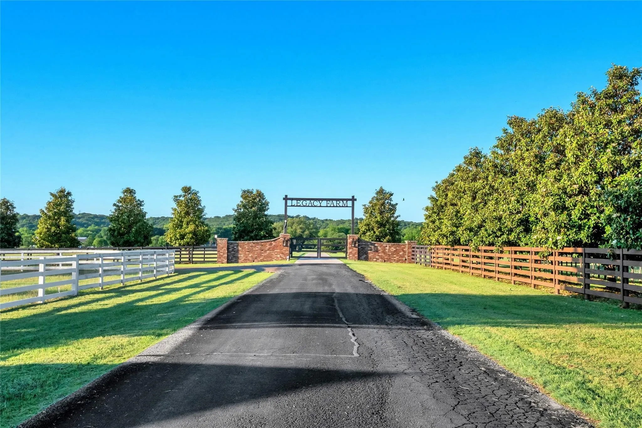 Paved driveway entrance with a gated entry, brick columns, white perimeter fencing, wood perimeter fencing, and mature trees