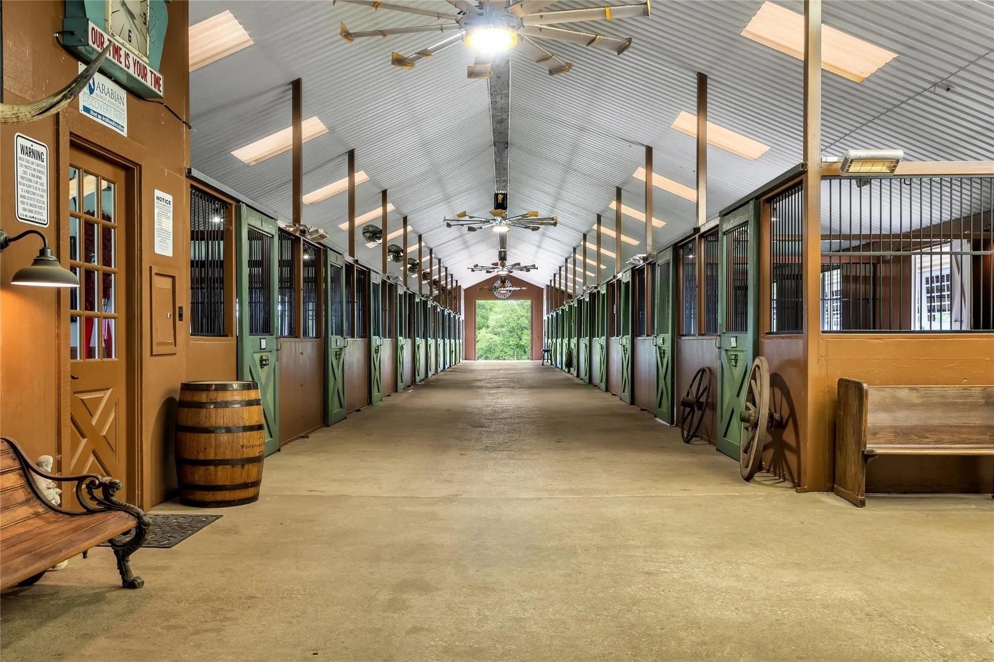 Expansive stable aisle featuring individual stalls with green wood-finish doors, exposed wood beams, and a corrugated metal ceiling