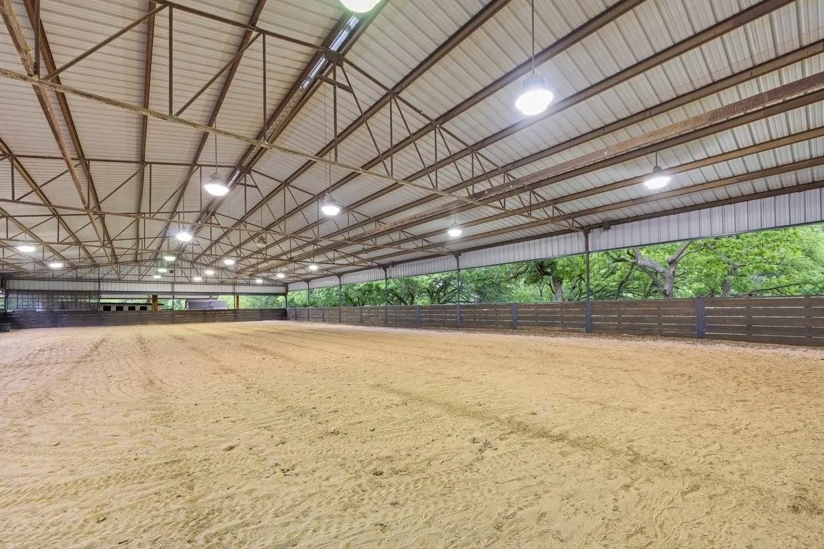 Spacious indoor equestrian arena featuring a sand-footed riding surface, expansive open layout with soaring vaulted ceiling and exposed steel beam construction, professional-grade LED lighting throughout, and large windowed perimeter allowing natural light and ventilation