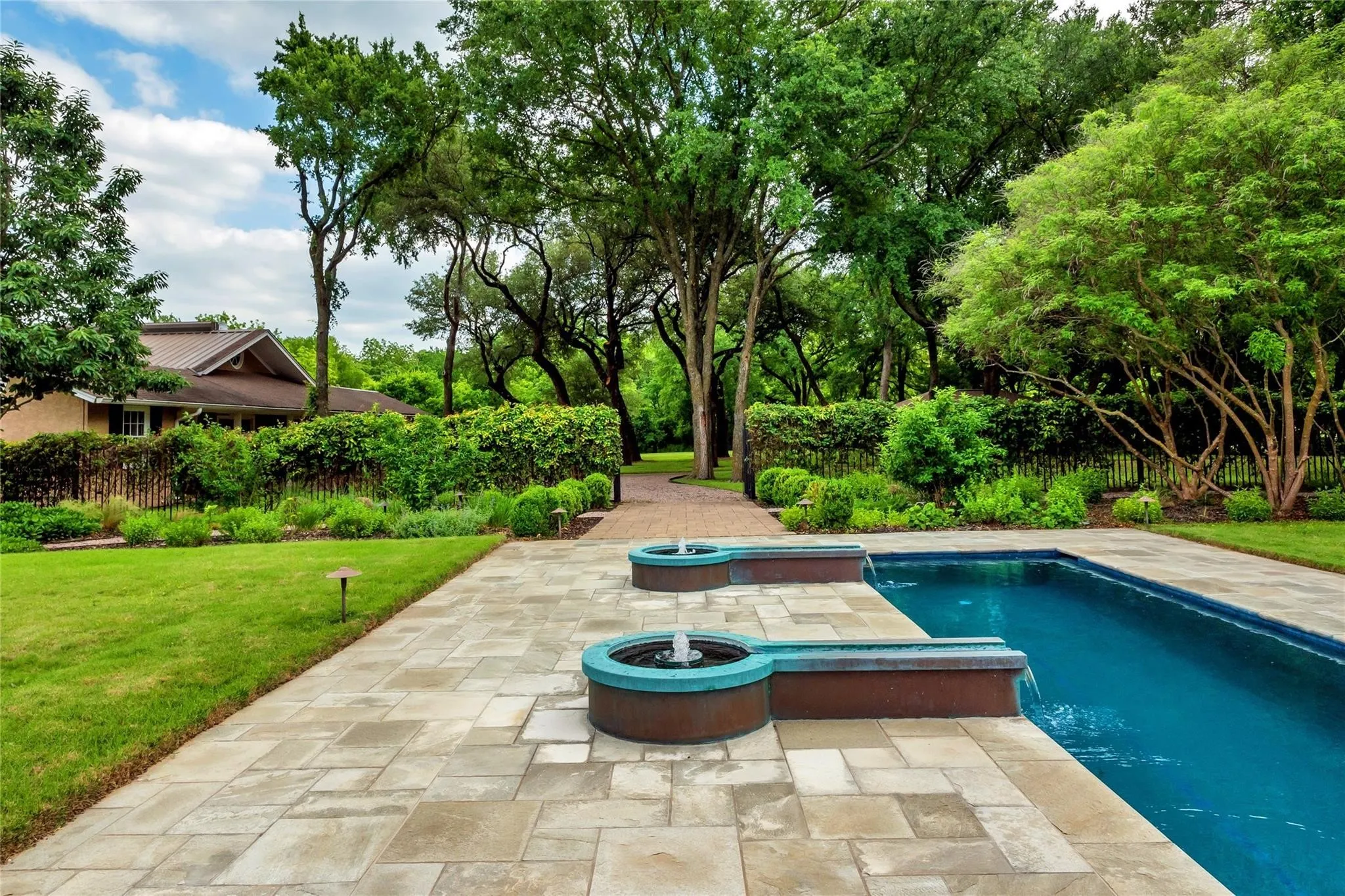 Swimming pool with water features, stone paver patio, lush landscaping, manicured lawn, and mature trees