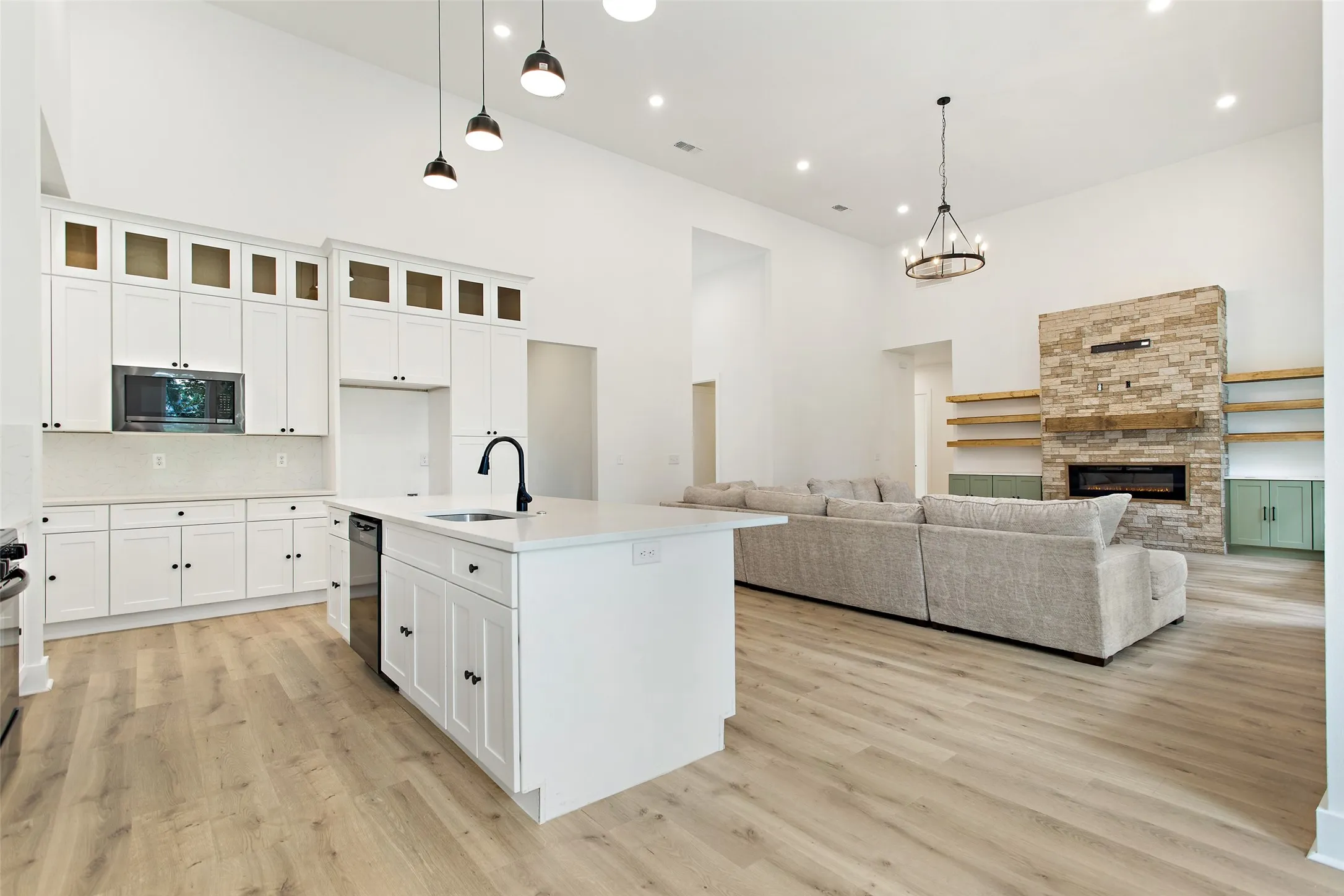 High vaulted ceiling with recessed lighting, wood-finish flooring, white cabinetry with display uppers, kitchen island with sink, and a stone-clad fireplace