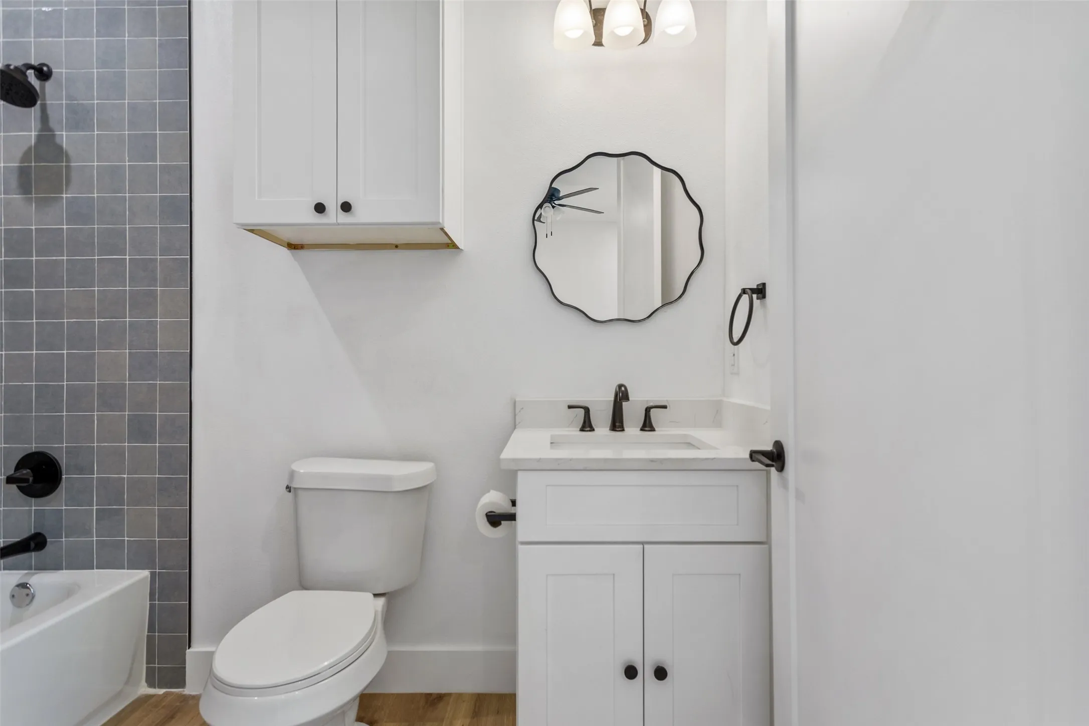 Modern bathroom featuring a white vanity with dual cabinet doors and an integrated sink, matte black hardware and faucet, a scalloped-edge mirror with black frame, white subway tile wall treatment, wood-finish flooring, and a white toilet with elongated bowl
