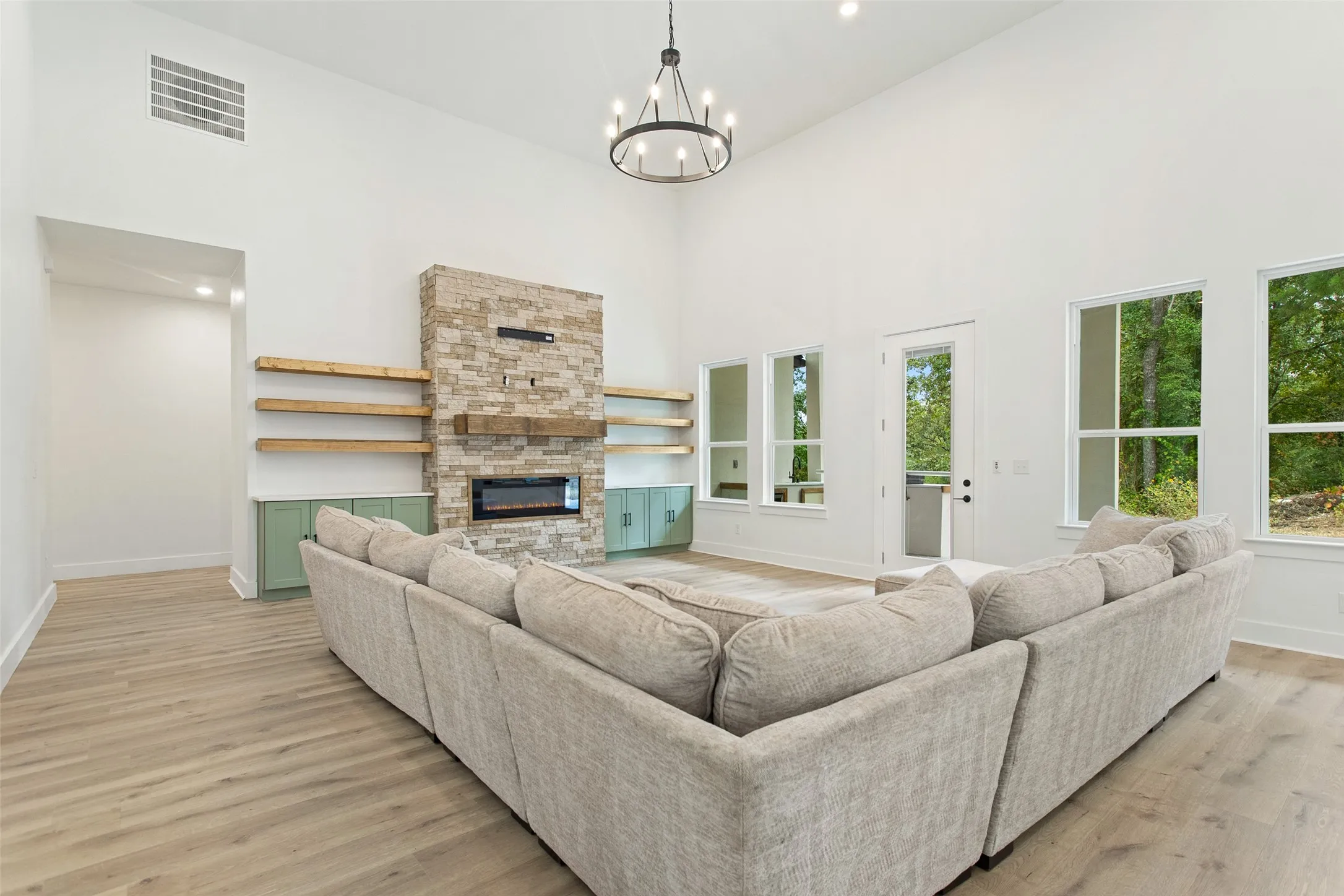 Vaulted ceiling with chandelier, stone-clad fireplace with mantel, built-in shelving, wood-finish flooring, and multiple windows