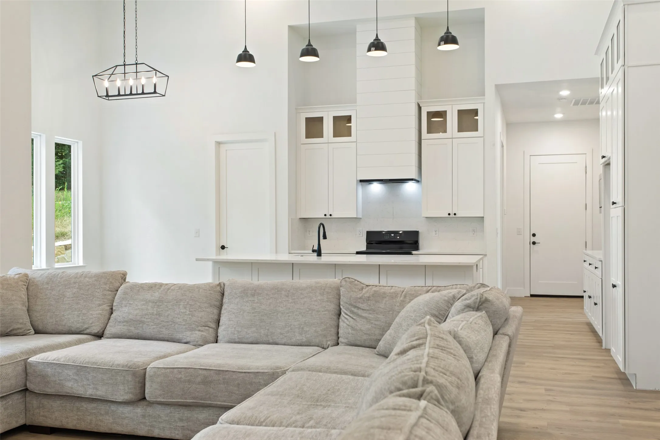 Open-concept living and kitchen area featuring a sectional seating arrangement, light wood-finish flooring throughout, custom white cabinetry with glass-front upper storage, stainless steel appliances, pendant lighting, and abundant natural light from large windows