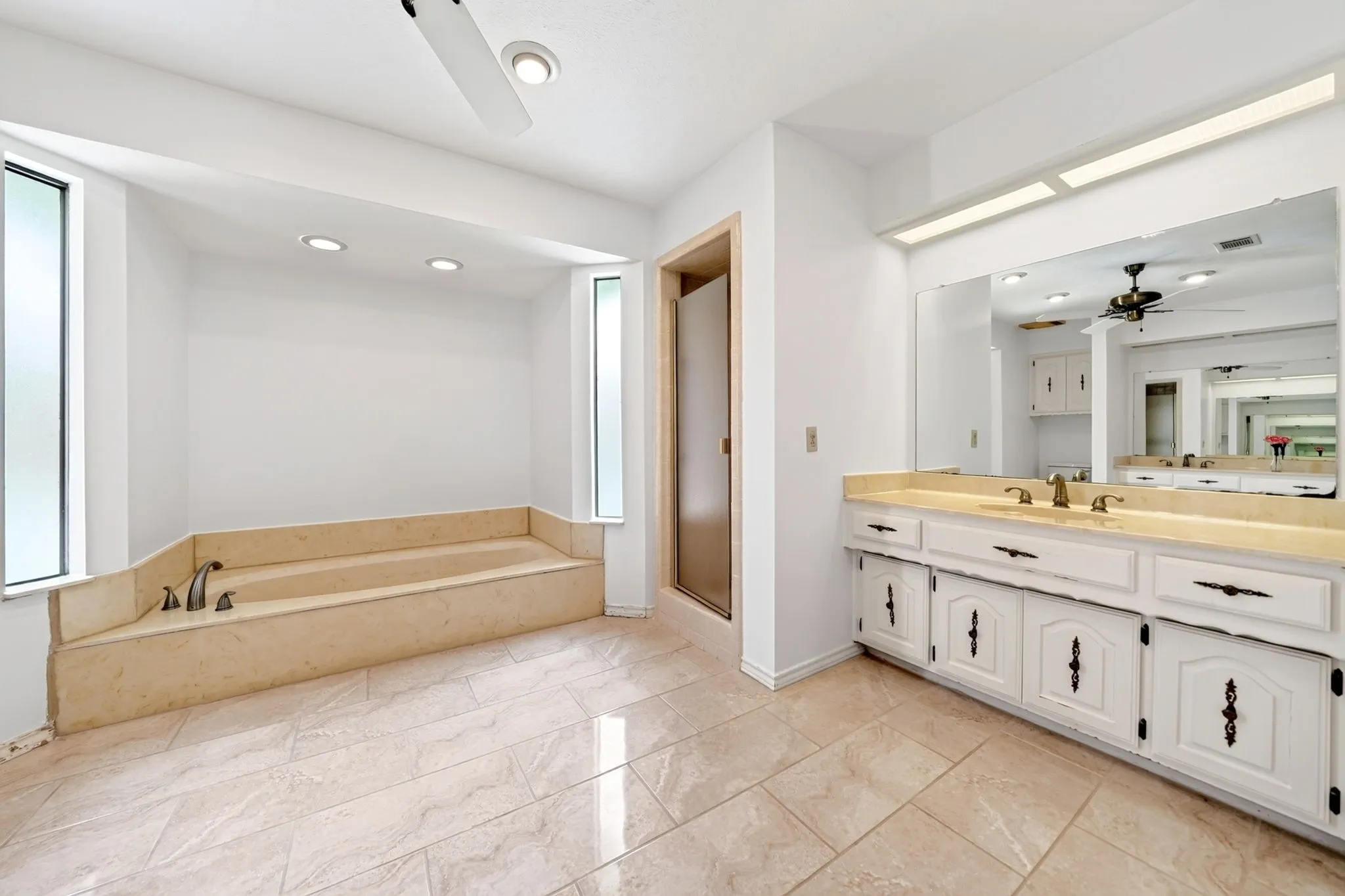 Spa-inspired primary bathroom featuring a soaking tub with wood-finish surround, dual-sink vanity with white cabinetry and black hardware, wood-finish countertop, large framed mirror, recessed and ceiling fan lighting, and neutral tile flooring throughout