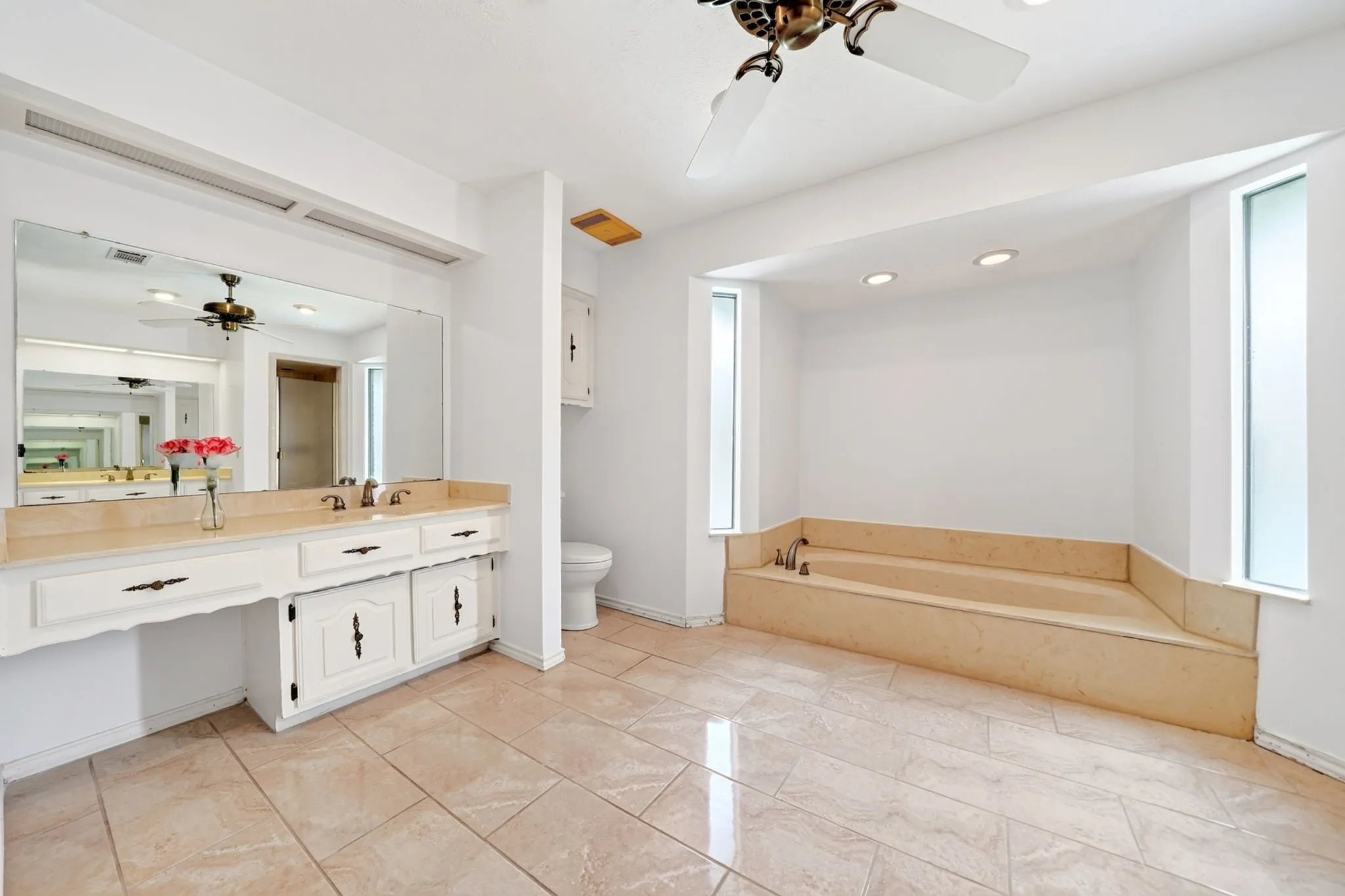 Spa-inspired primary bathroom featuring dual-sink vanity with white cabinetry and wood-finish countertop, large soaking tub with tile surround, tile flooring throughout, abundant natural light from multiple windows, and recessed ceiling lighting