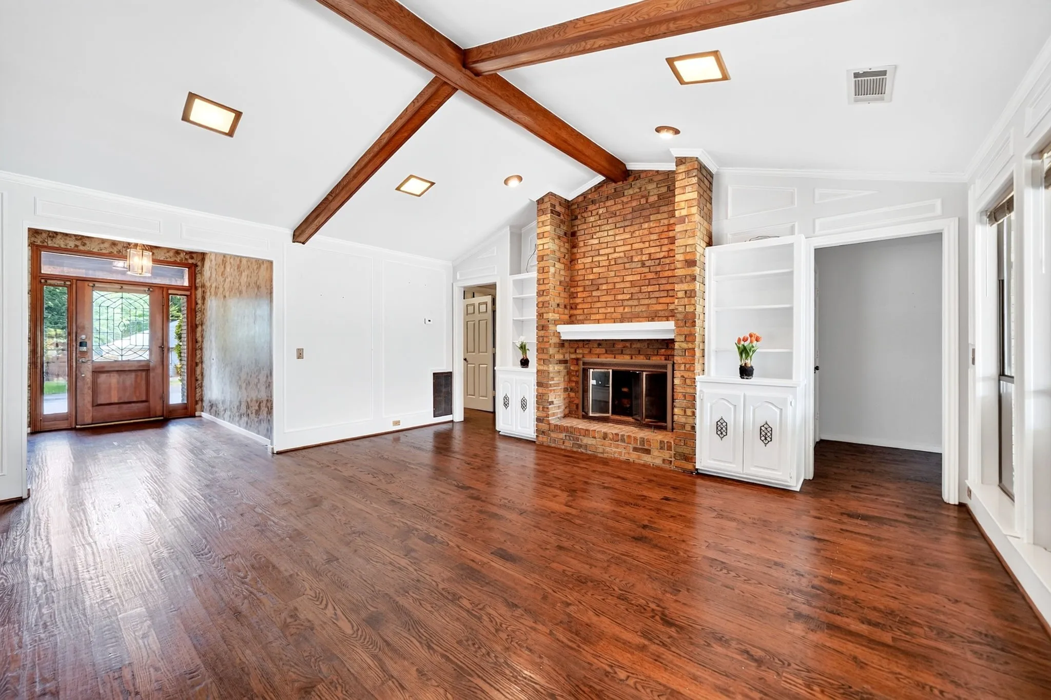 Vaulted ceiling with wood beams, brick fireplace with white mantel, wood-finish flooring, built-in cabinetry, and a wood-paneled entry door with decorative glass