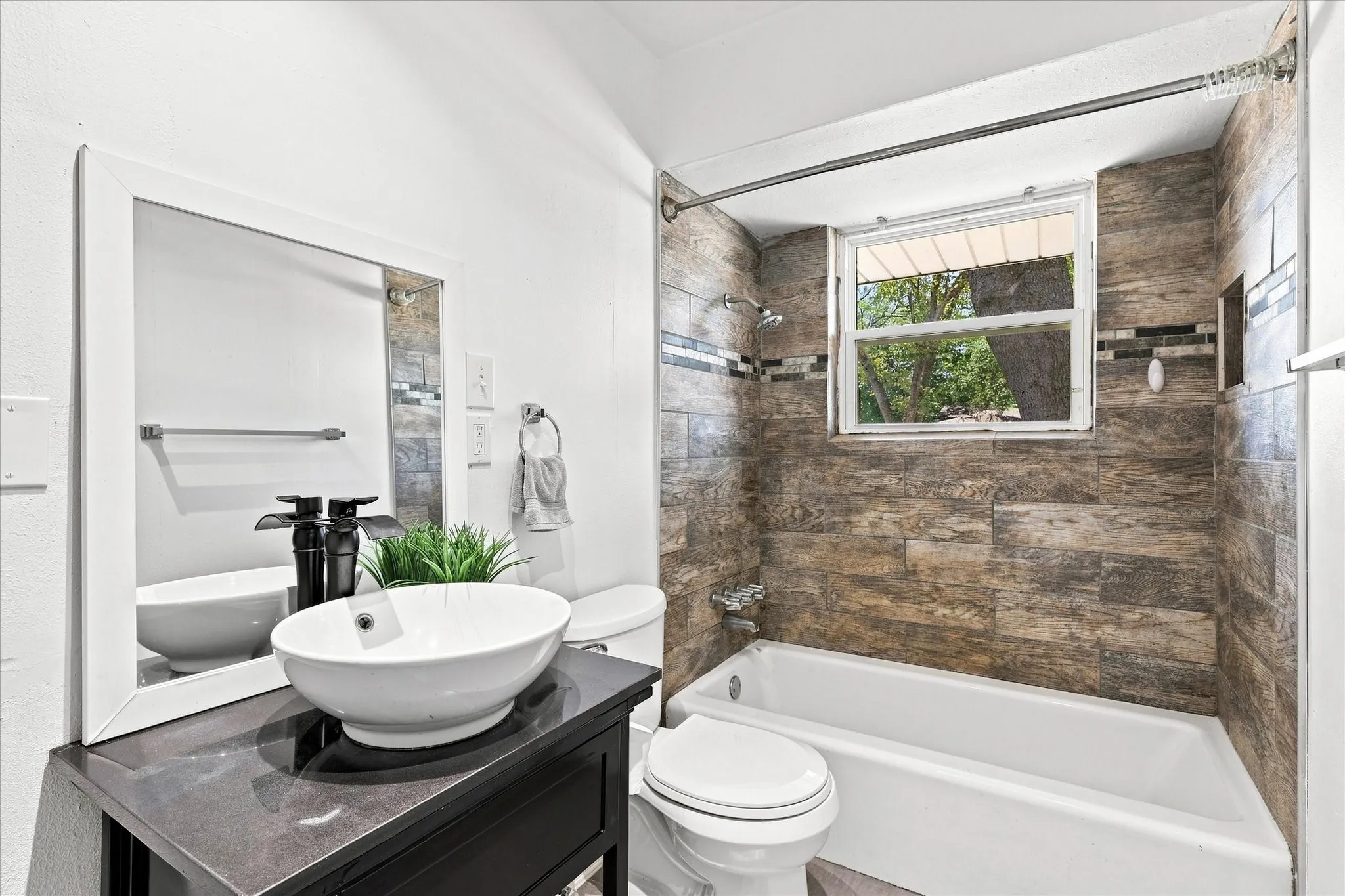 Bathroom featuring a vessel sink, dark wood-finish vanity, white framed mirror, and a tiled shower-tub combination with an integrated window