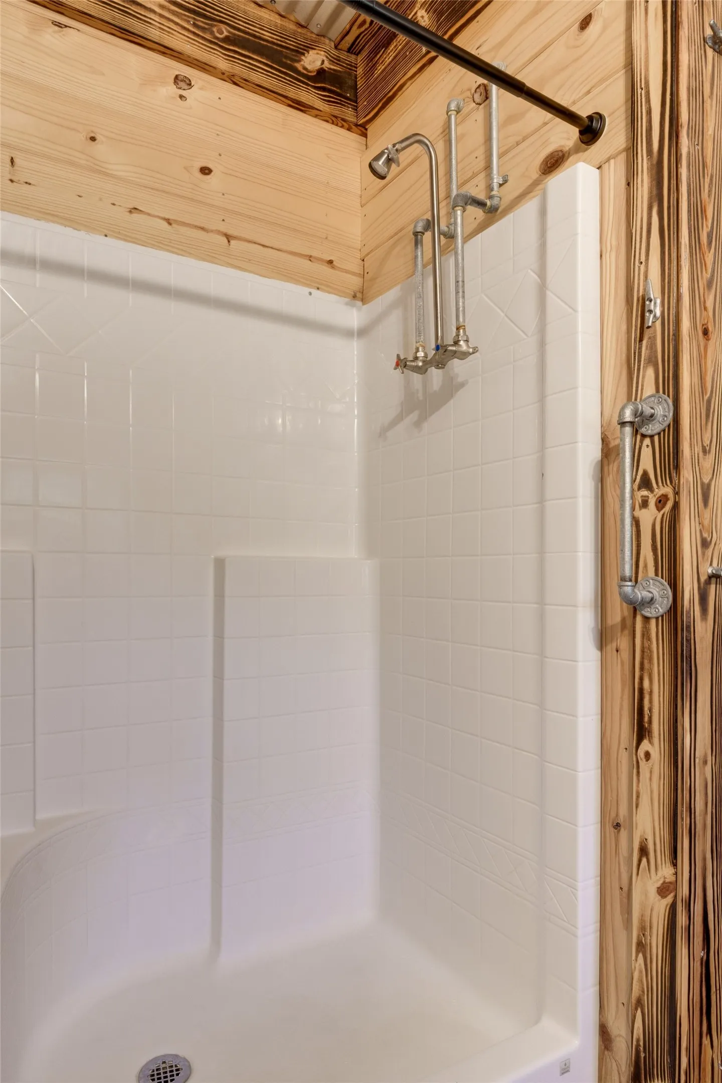 White shower stall with a grid pattern, industrial-style exposed pipe shower fixtures, and a wood-paneled wall