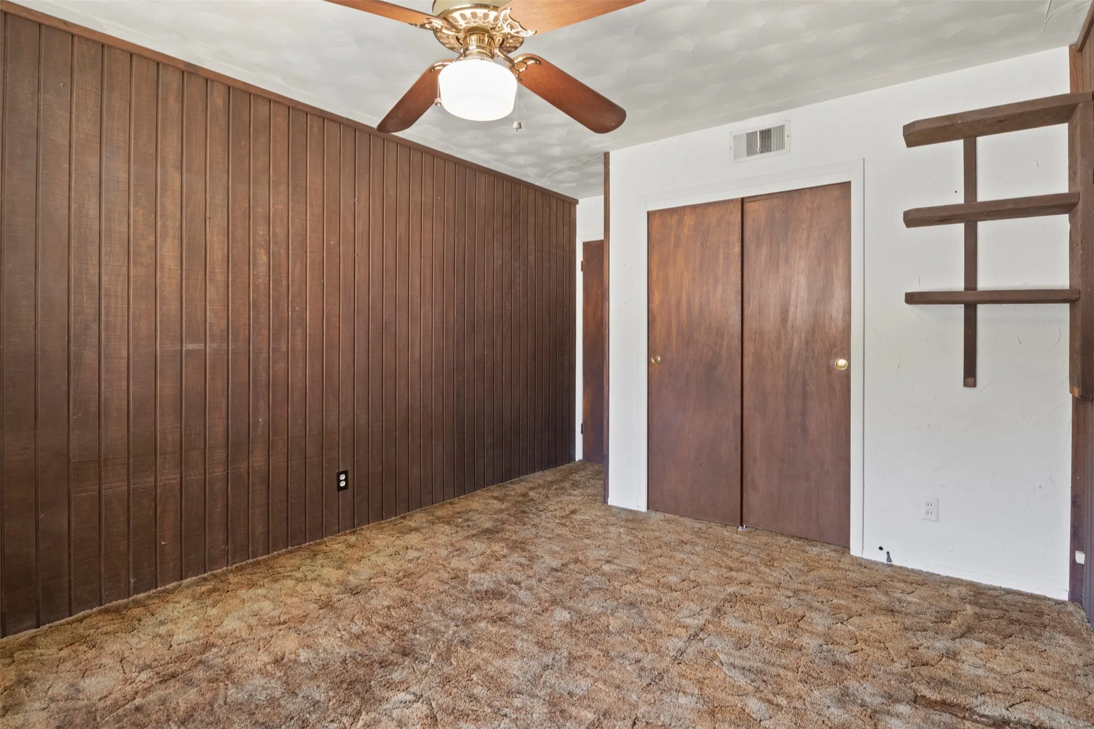 Wood paneling accent wall, ceiling fan with integrated lighting, wood-finish bi-fold closet doors, and built-in shelving
