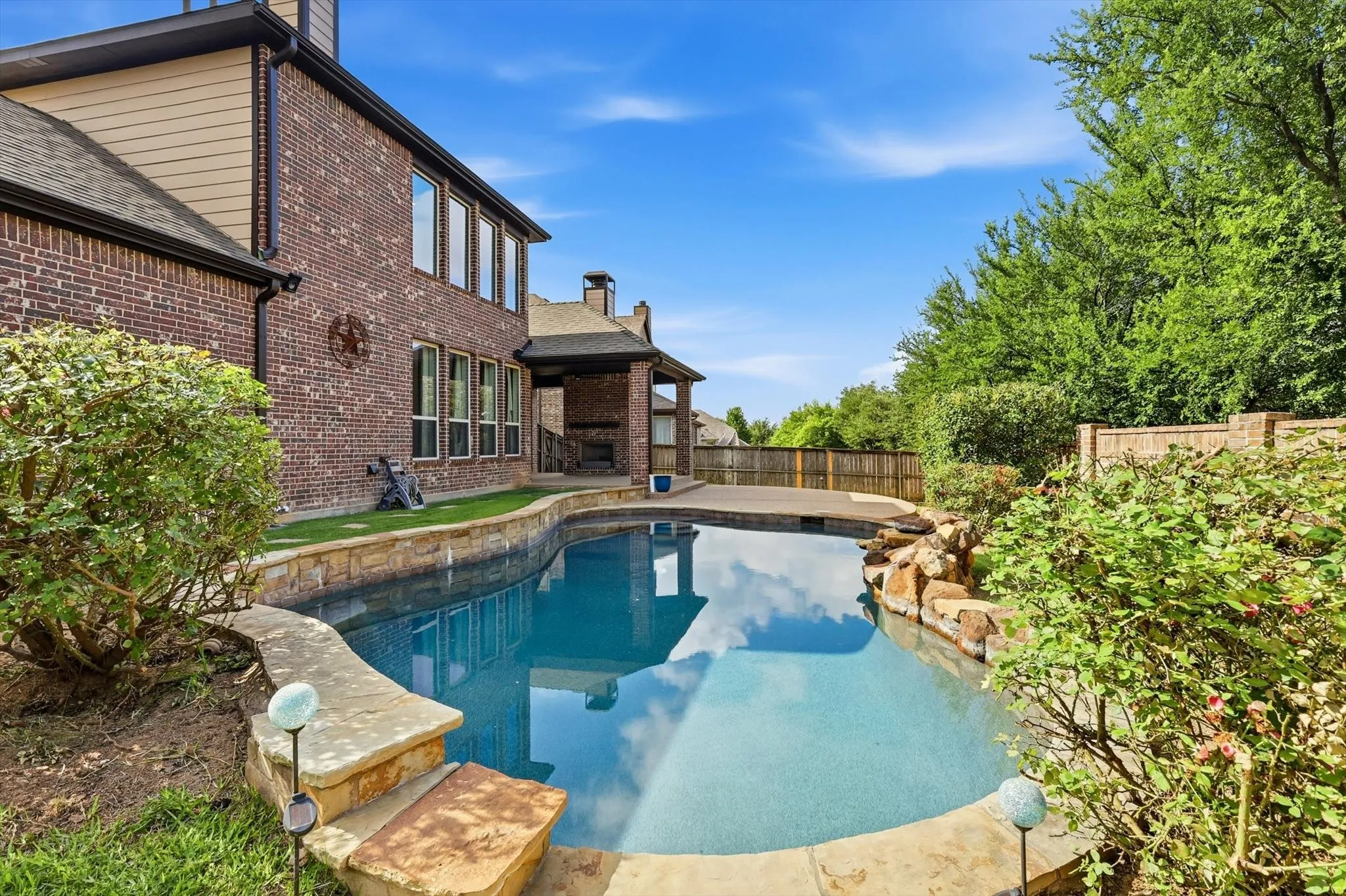 Curved swimming pool with stone coping, a rock water feature, a covered patio, and a brick exterior