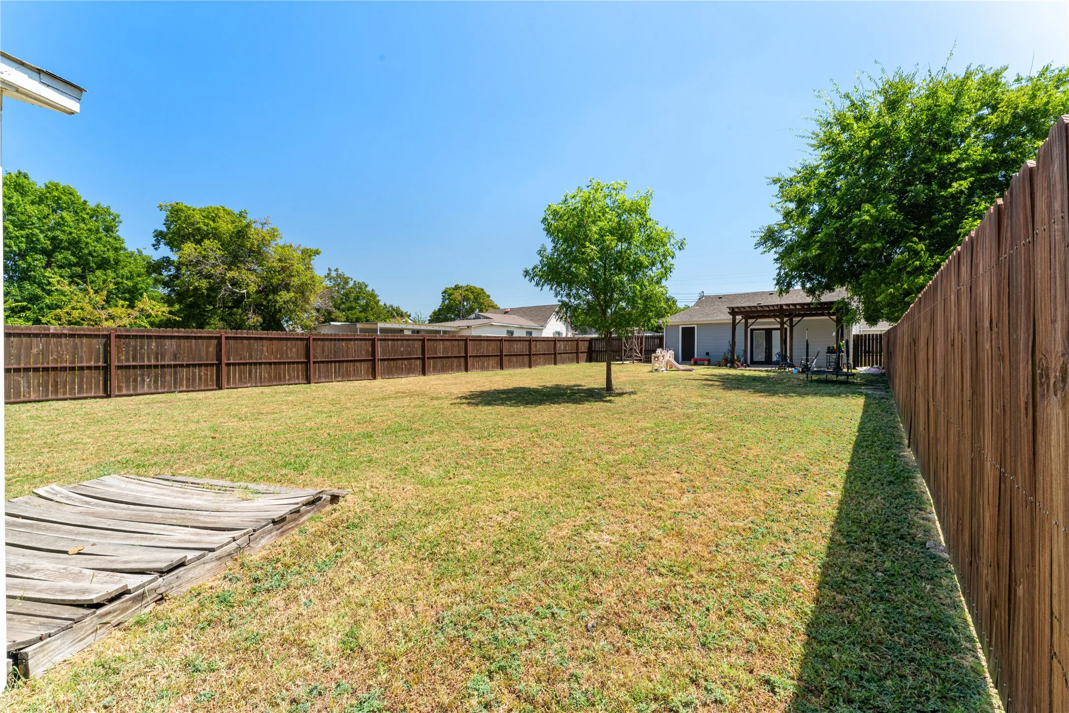 Expansive backyard featuring a stained wood privacy fence, a mature shade tree, and a pergola-covered patio