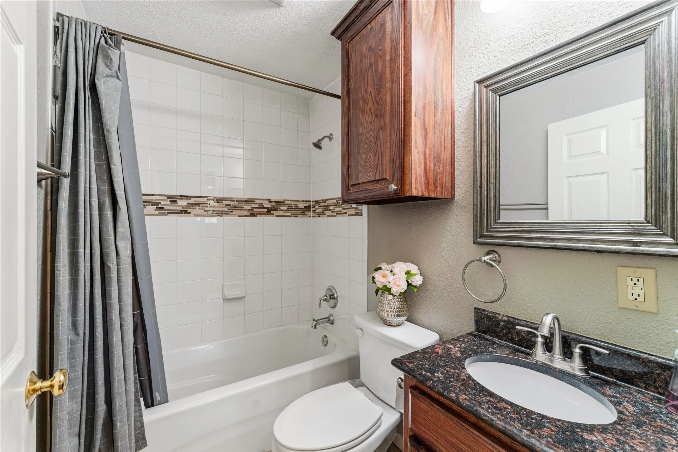 Built-in tub with tile surround and decorative accent band, dark wood-finish vanity with stone countertop and integrated sink, framed wall mirror, and wall-mounted storage cabinet
