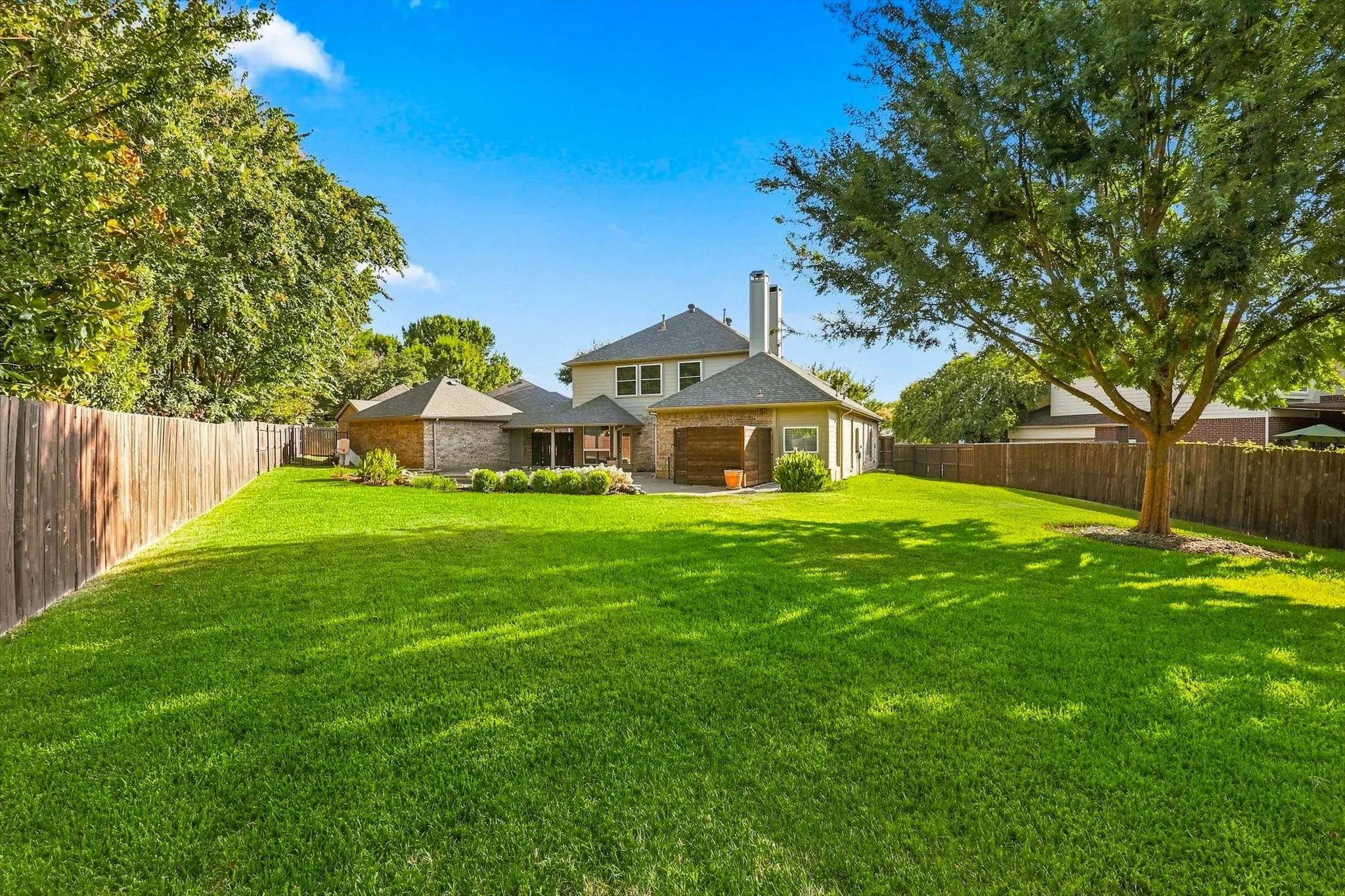 Expansive manicured lawn with mature trees, a wood privacy fence, and a brick exterior home with a covered patio