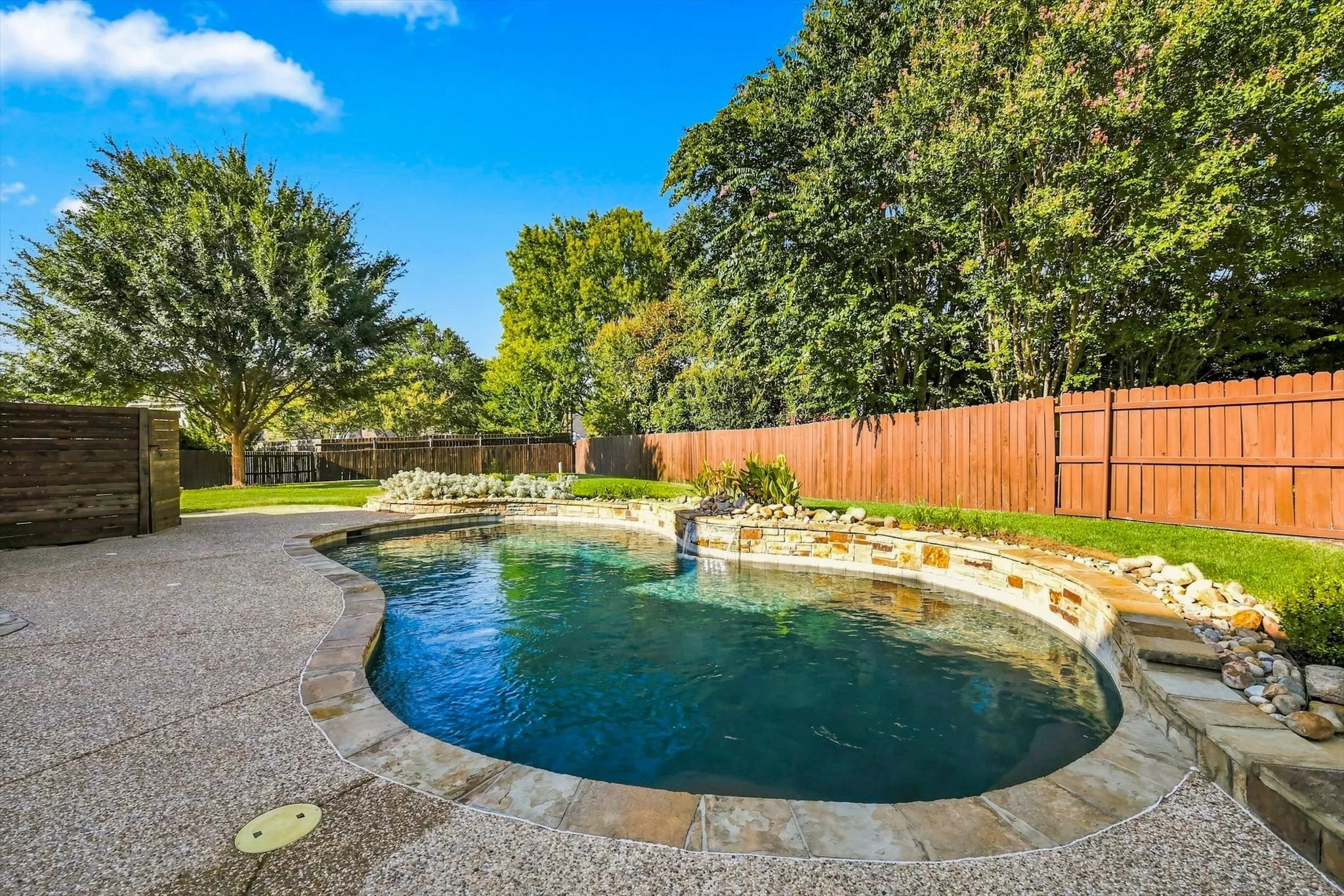 Freeform swimming pool with stone coping, natural rock waterfall, and integrated landscaping, surrounded by an aggregate concrete deck and wood privacy fencing
