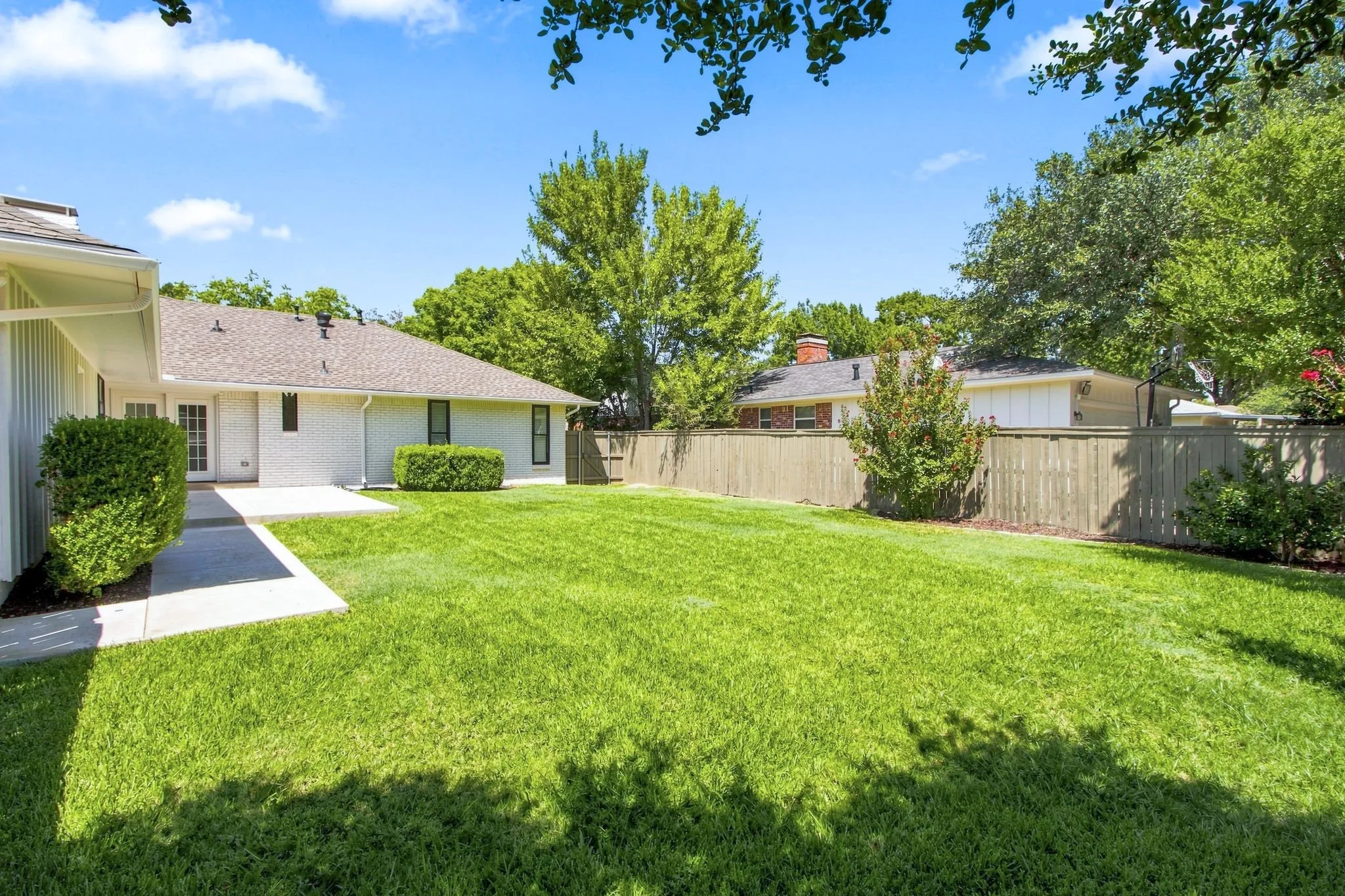 Expansive green lawn, concrete patio, mature perimeter trees, wood fence, and a white brick exterior