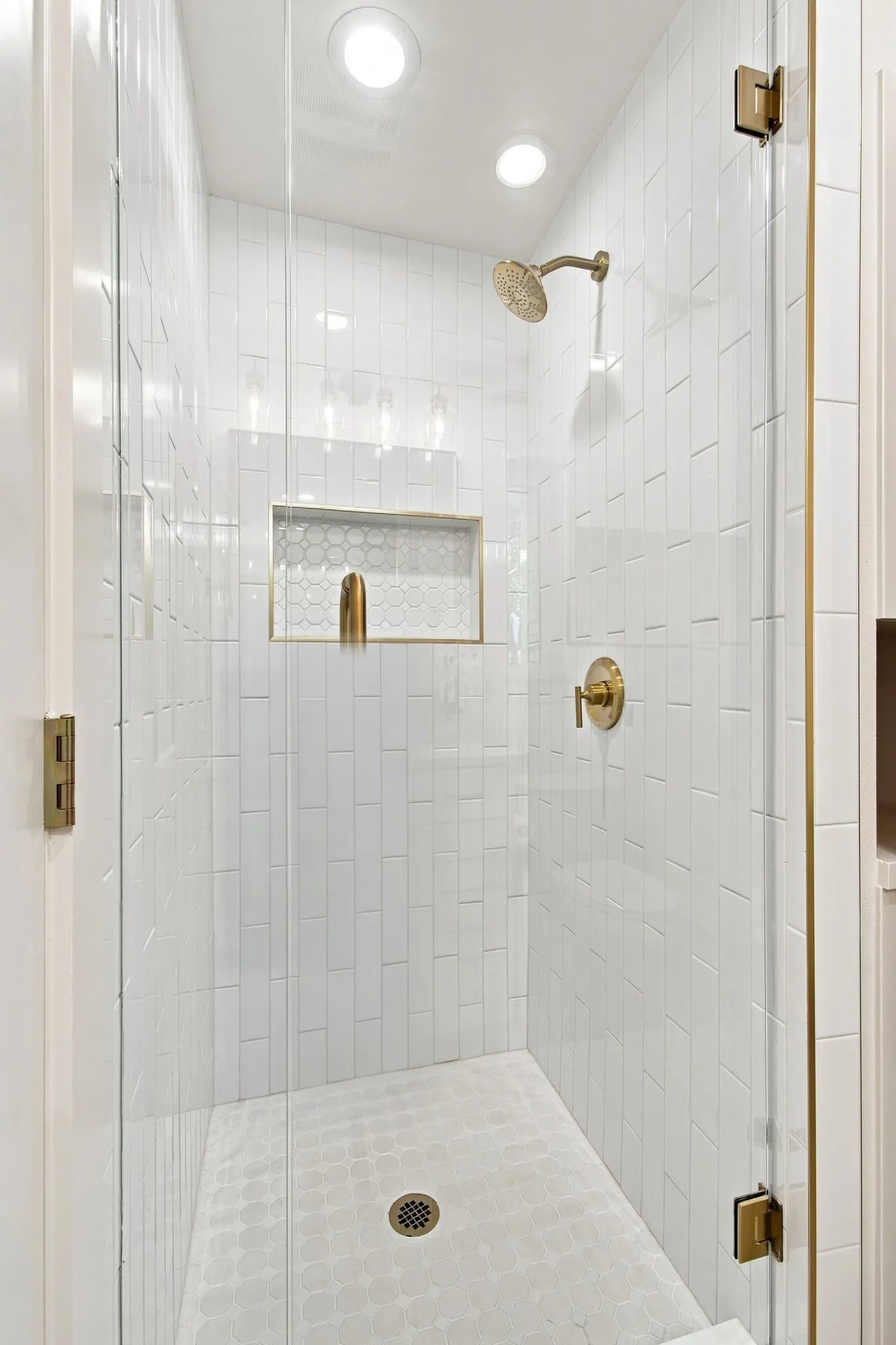 Walk-in shower with clear glass enclosure and brass hardware, vertical subway tile surround, recessed niche with hexagon tile, and a circular tile shower floor