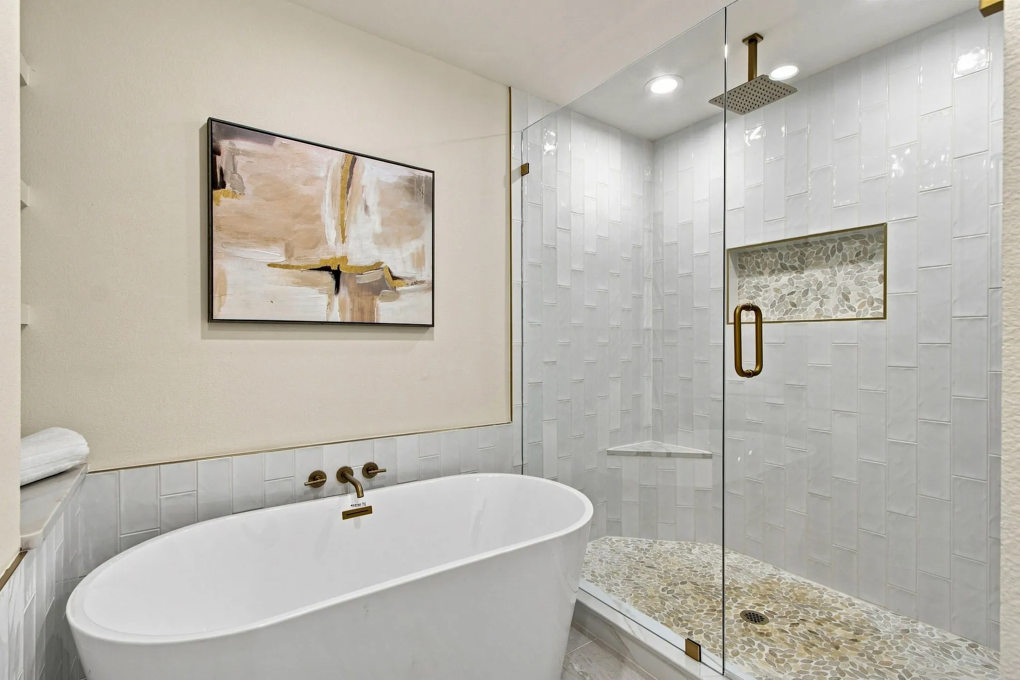 Freestanding soaking tub with wall-mounted bronze faucet, glass-enclosed shower with subway tile surround, recessed shower niche, and pebble-finish flooring, complemented by a ceiling-mounted bronze rain shower head