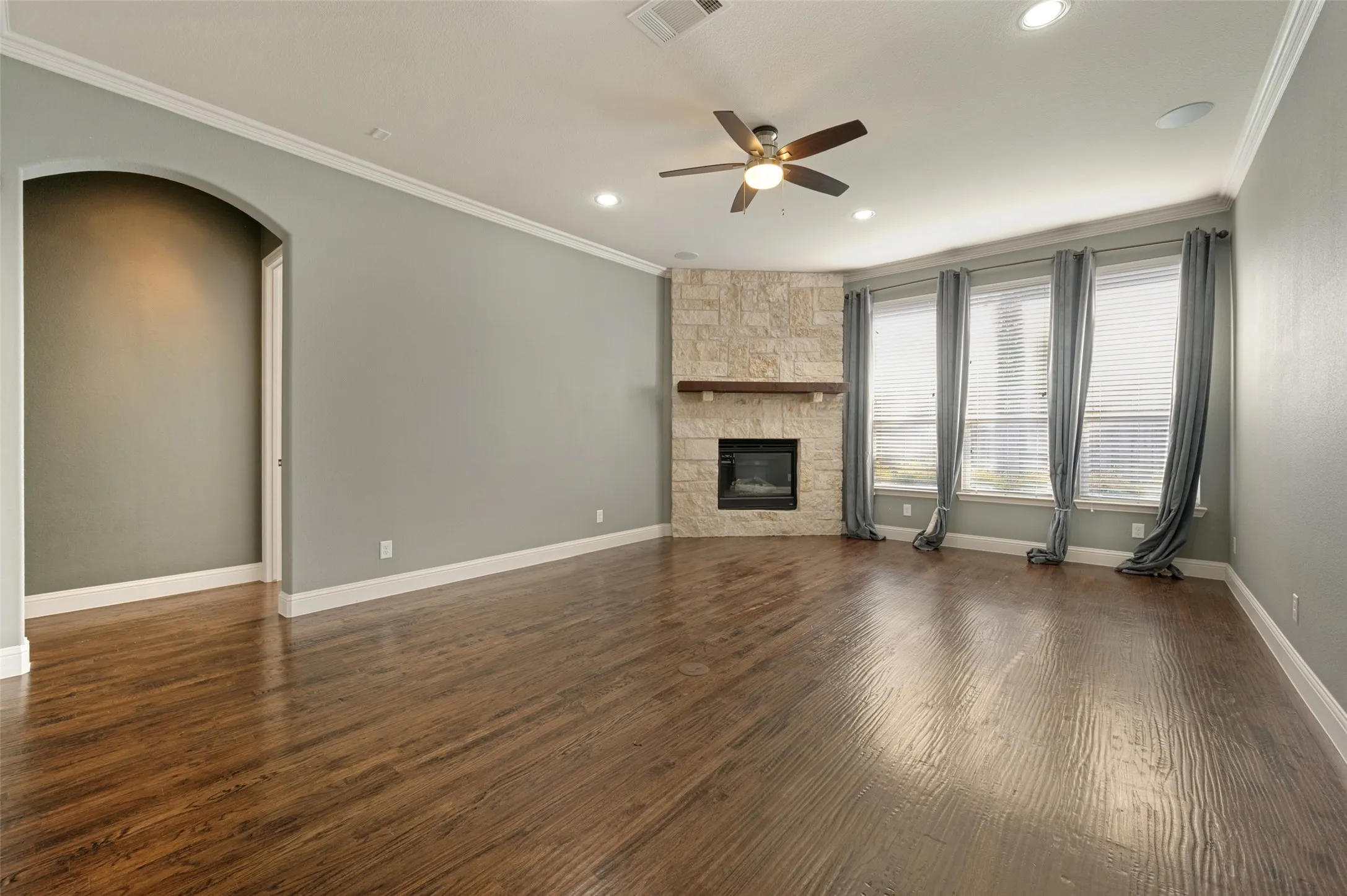 Wood-finish flooring, recessed lighting, crown molding, a stone-clad fireplace, and multiple windows with blinds
