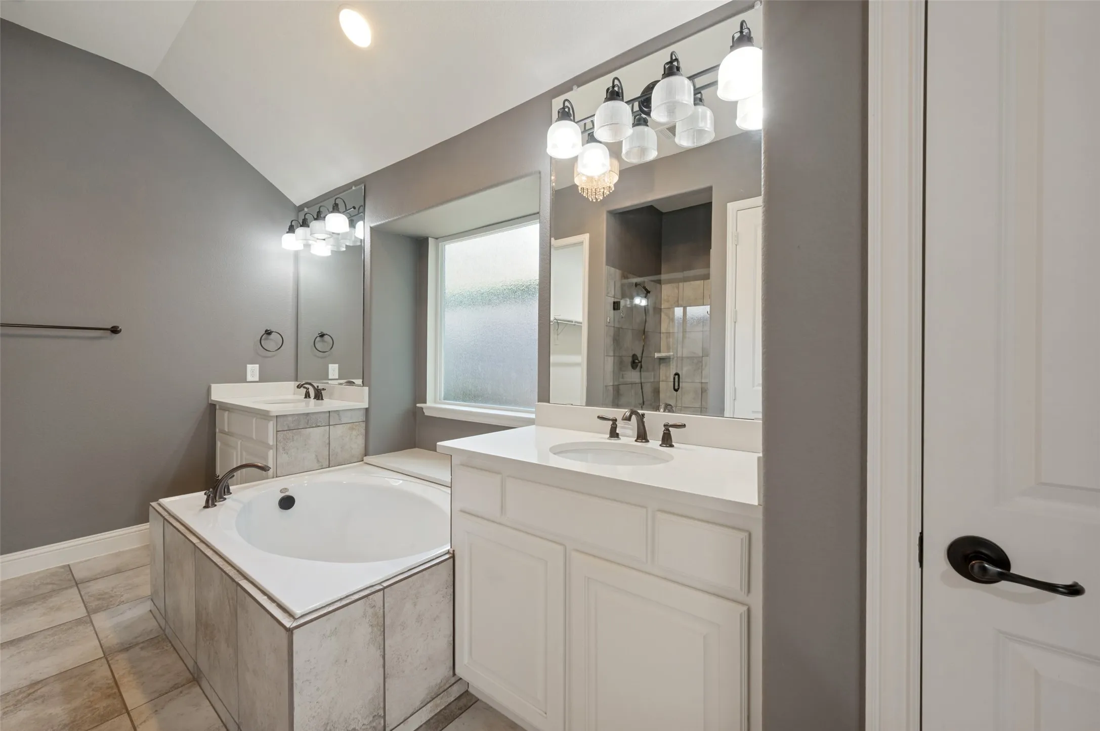 Vaulted ceiling with recessed lighting, two white vanities with undermount sinks, built-in bathtub with tiled surround, and tiled flooring