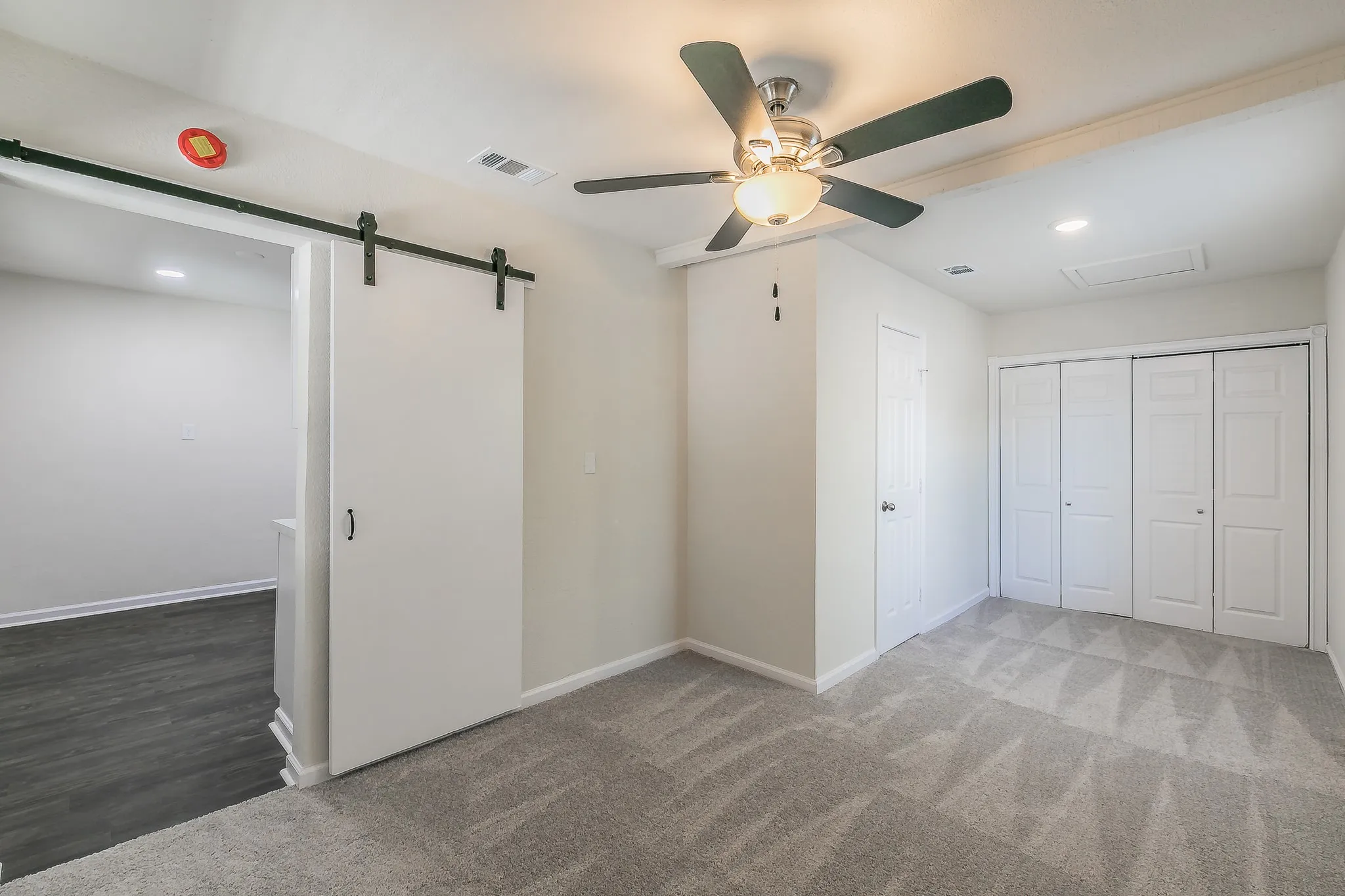 Gray patterned carpeting, white barn door on black hardware, ceiling fan with light, recessed lighting, and white bi-fold closet doors