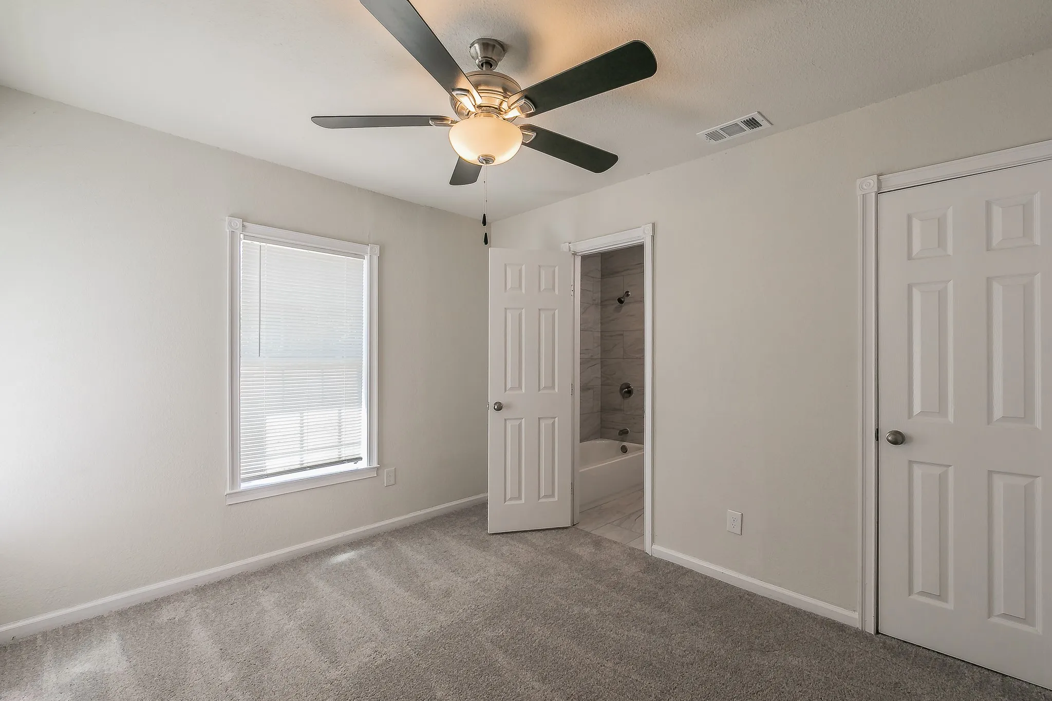 Carpeted room with a ceiling fan, a window with blinds, and an open doorway revealing a bathroom with a tiled shower and bathtub
