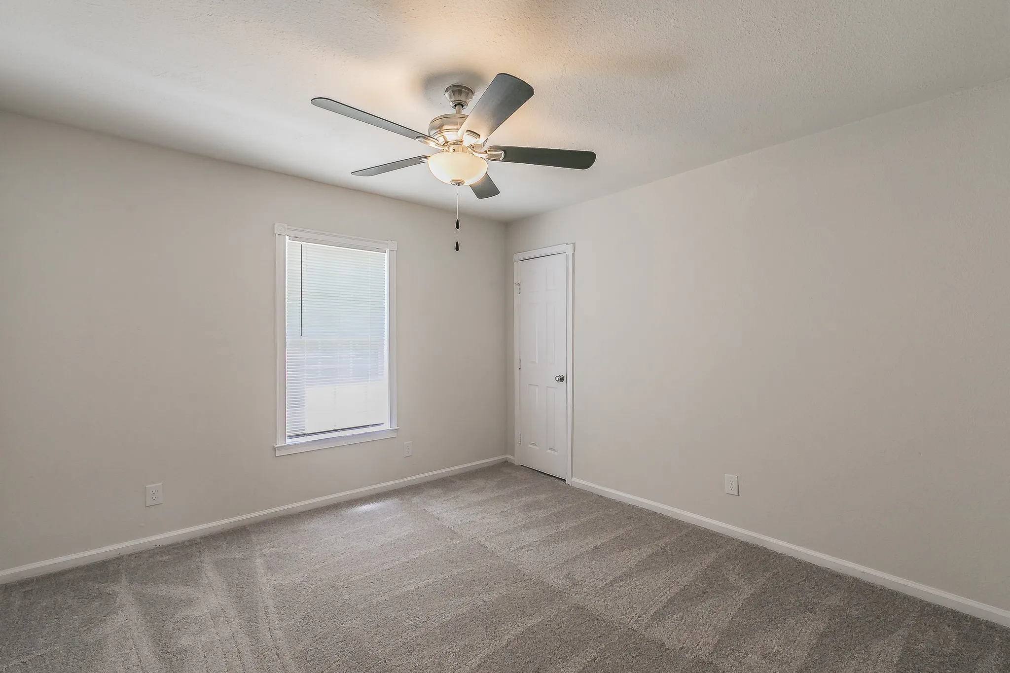 Light gray wall paint, textured ceiling, gray carpet flooring, window with blinds, and a ceiling fan with light fixture